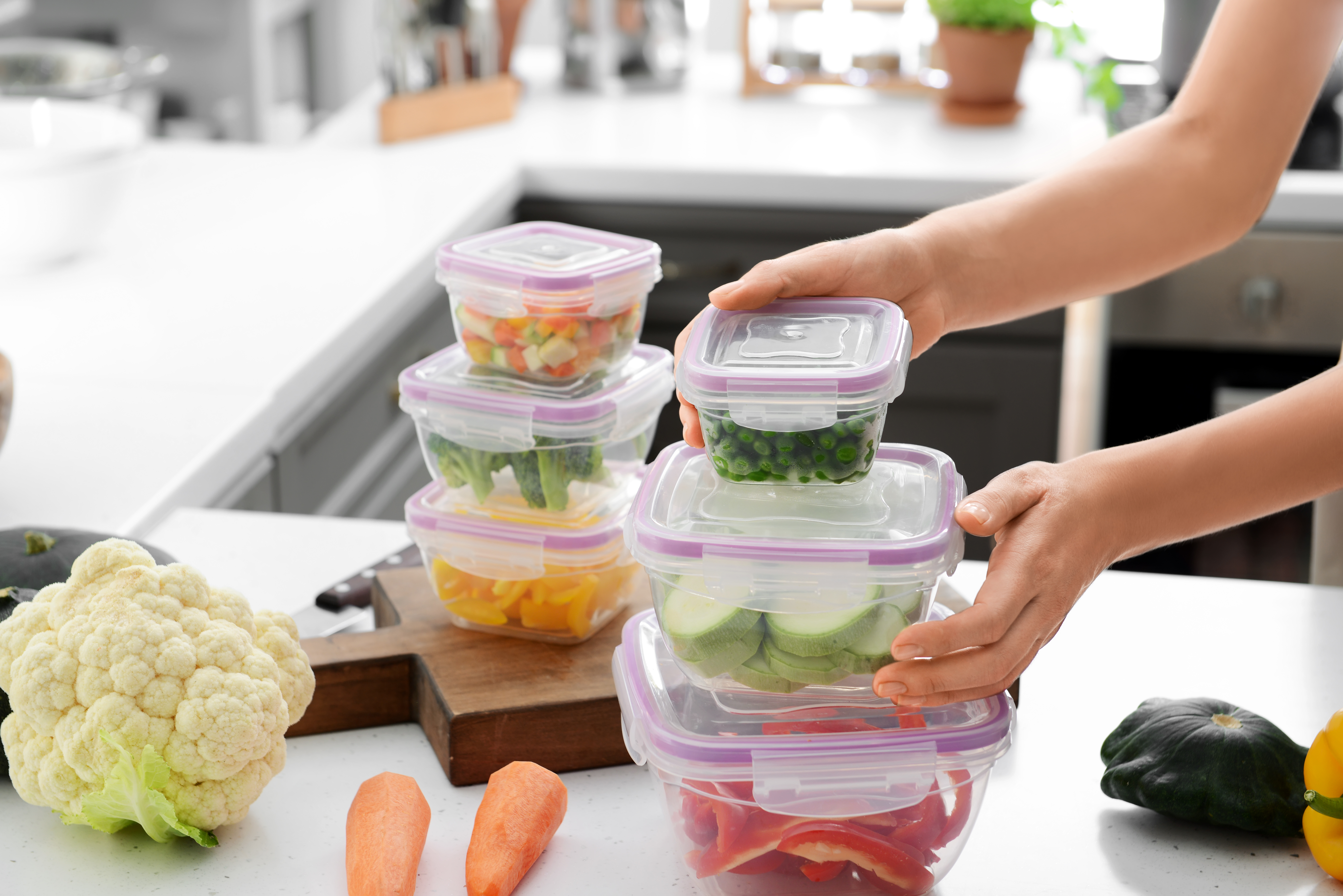 Femme tenant une pile de récipients en plastique contenant des légumes frais à congeler à une table dans la cuisine | Source : Shutterstock