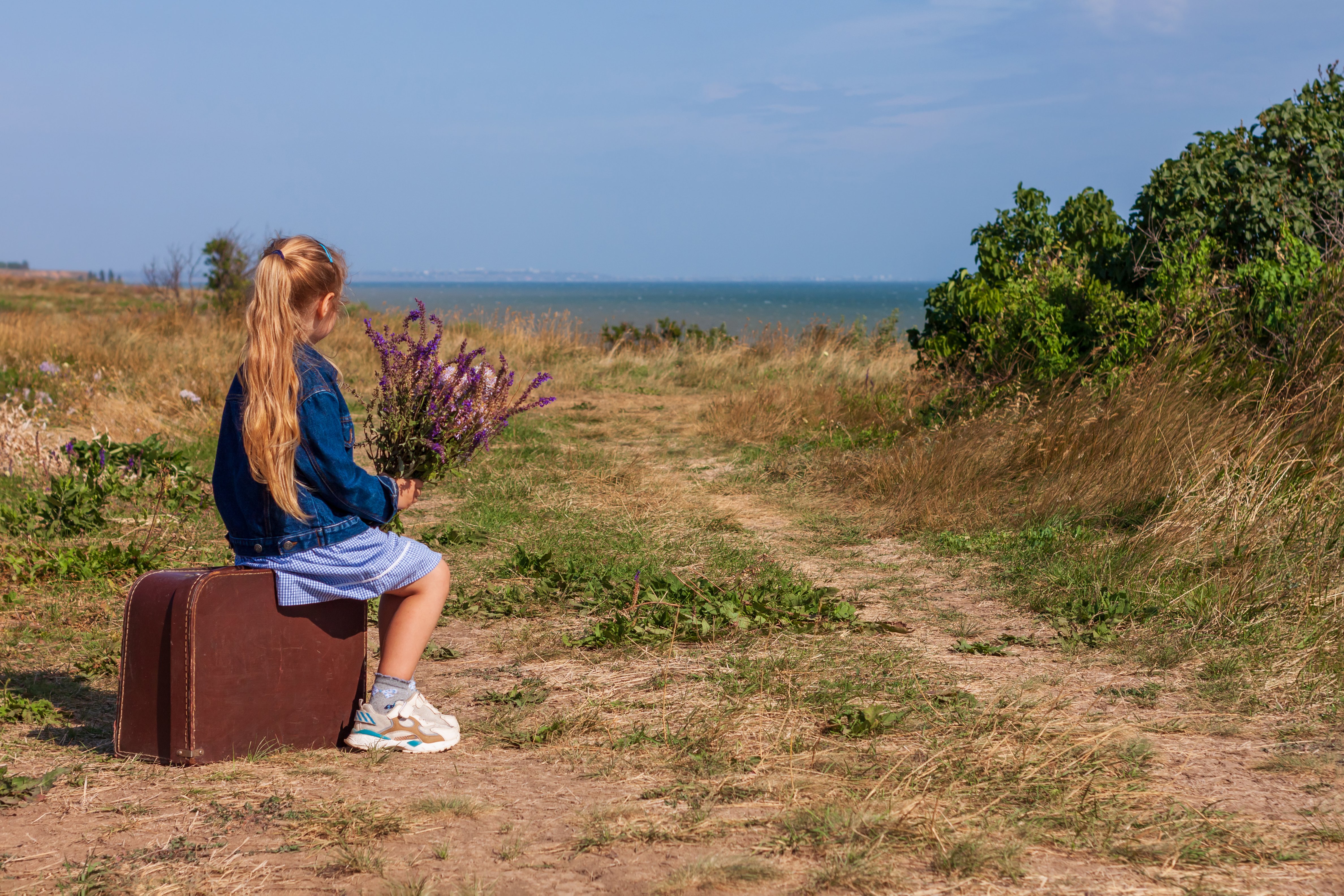 Petite fille blonde en veste de denim, robe bleue avec valise vintage et bouquet de fleurs hors route avec paysage de mer. | Photo : Shutterstock