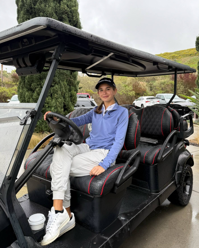 Kai est assis au volant d'une voiturette de golf, vêtu d'un haut bleu et d'un pantalon blanc, souriant avec une tasse de café à proximité | Source : Facebook/Kai-Trump