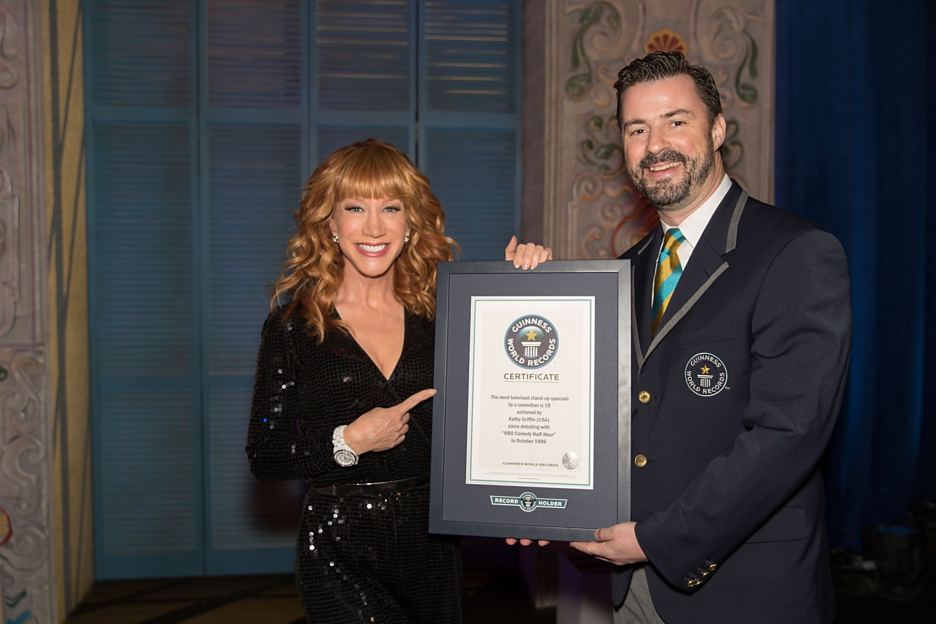 Kathy Griffin avec un représentant du Guinness World Record après avoir battu le record du monde Guinness du plus grand nombre d'émissions spéciales de stand-up par un humoriste, le 24 novembre 2013. | Source : Getty Images
