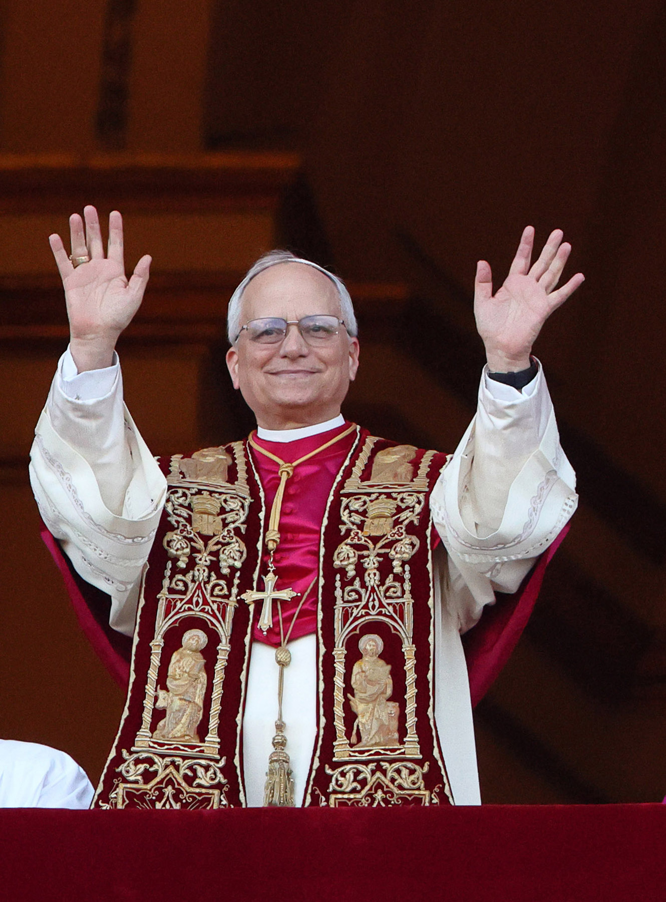 Le pape Léon XIV sourit chaleureusement en levant les deux mains en guise de salut, son étole cramoisie ornée de saints dorés captant la lumière