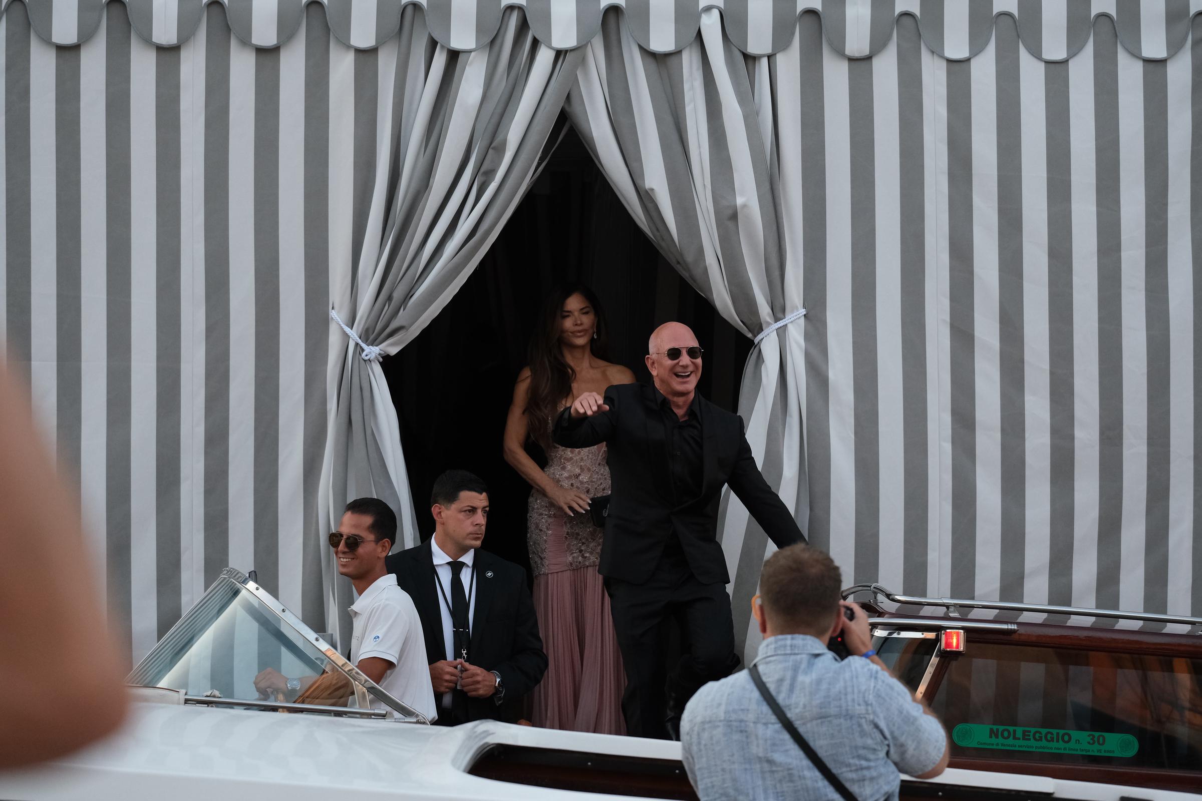 Jeff Bezos sourit et salue avec enthousiasme en montant à bord d'un bateau-taxi, suivi de Lauren Sánchez vêtue d'une robe à perles couleur rose pâle, encadrée par des rideaux rayés et entourée de membres du personnel et de badauds.