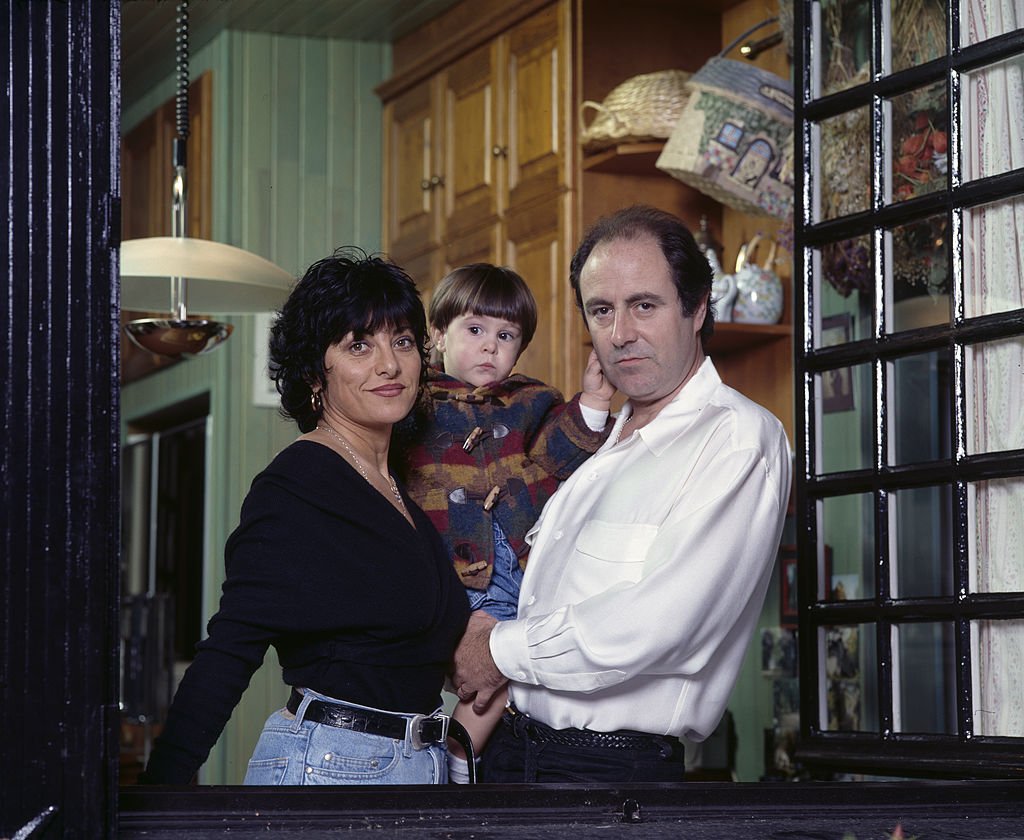 Dans l'embrasure d'une fenêtre de sa maison, portrait du chanteur Michel DELPECH tenant son fils Emmanuel DELPECH dans ses bras, en compagnie de son épouse Geneviève DELPECH | Photo : Getty Images.
