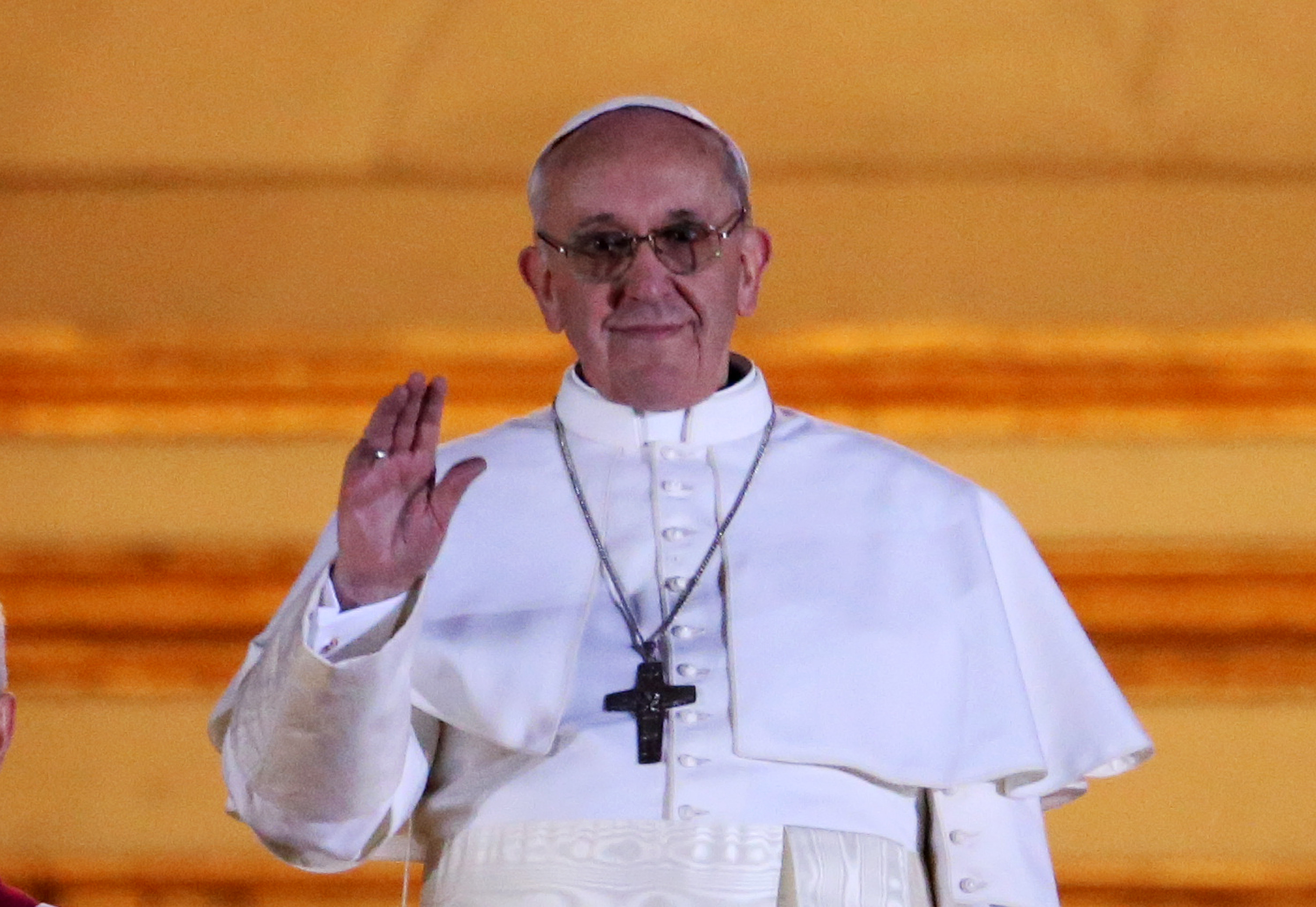 Le pape François salue la foule d'un léger signe de la main, vêtu d'une simple robe blanche et d'une croix argentée, son sourire calme encadré par une douce lumière dorée