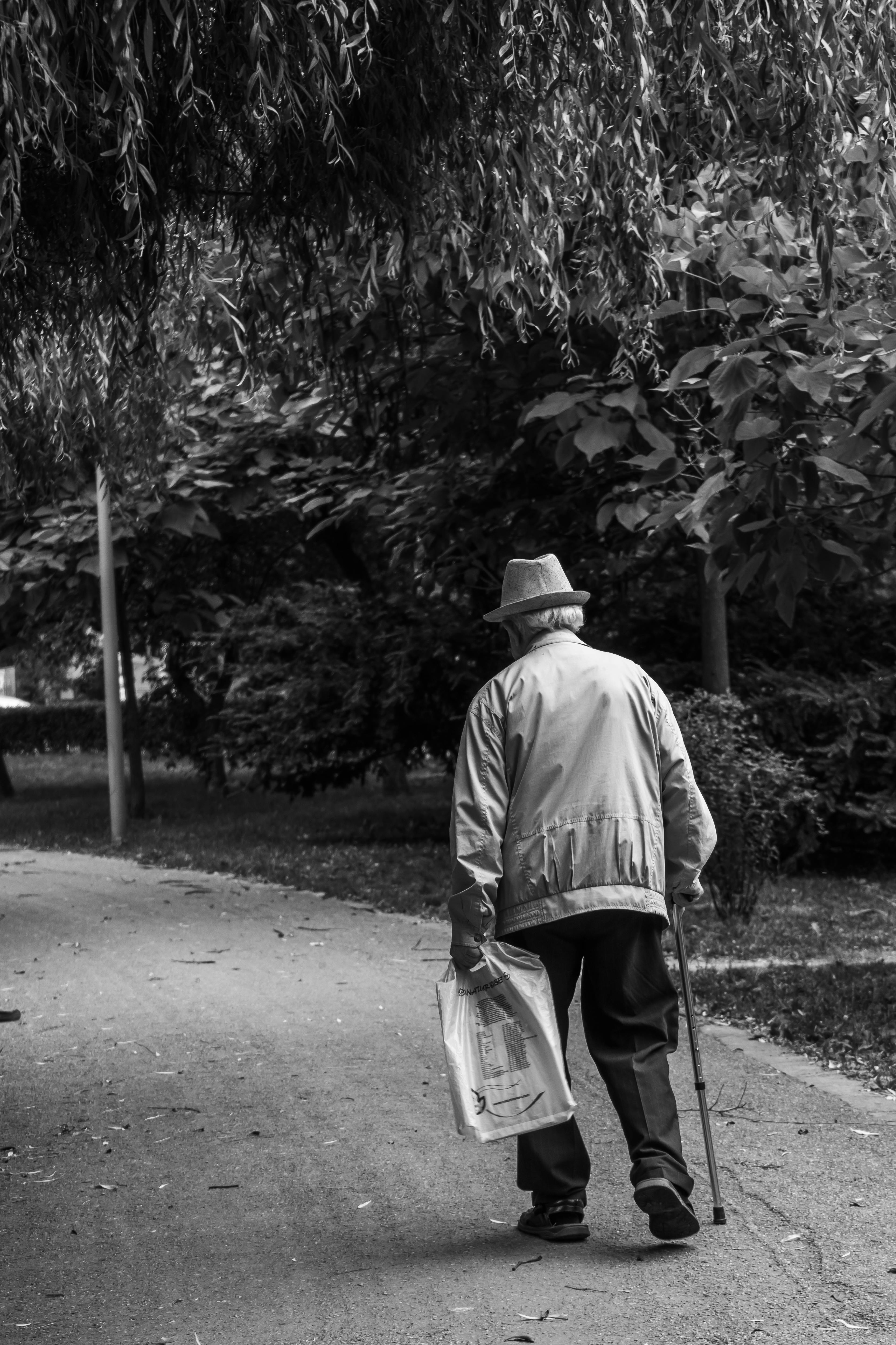 Niveaux de gris d'un vieil homme marchant avec une canne et un journal | Source : Pexels