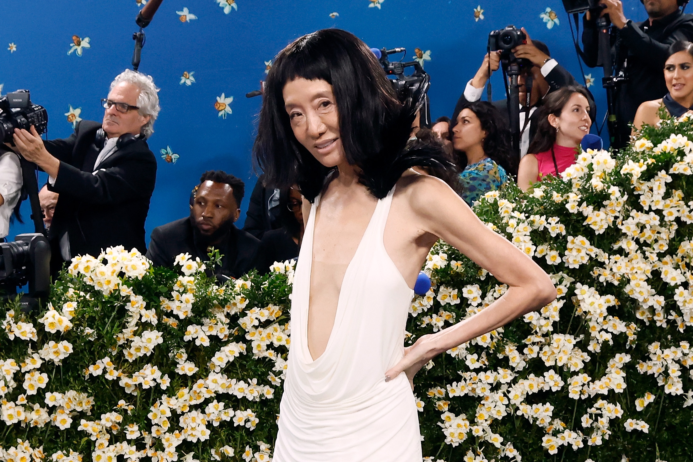 Prenant une pose assurée, Vera Wang se tient devant un champ de fleurs blanches, mettant en valeur le décolleté plongeant et les épaules plumeuses de sa robe personnalisée.