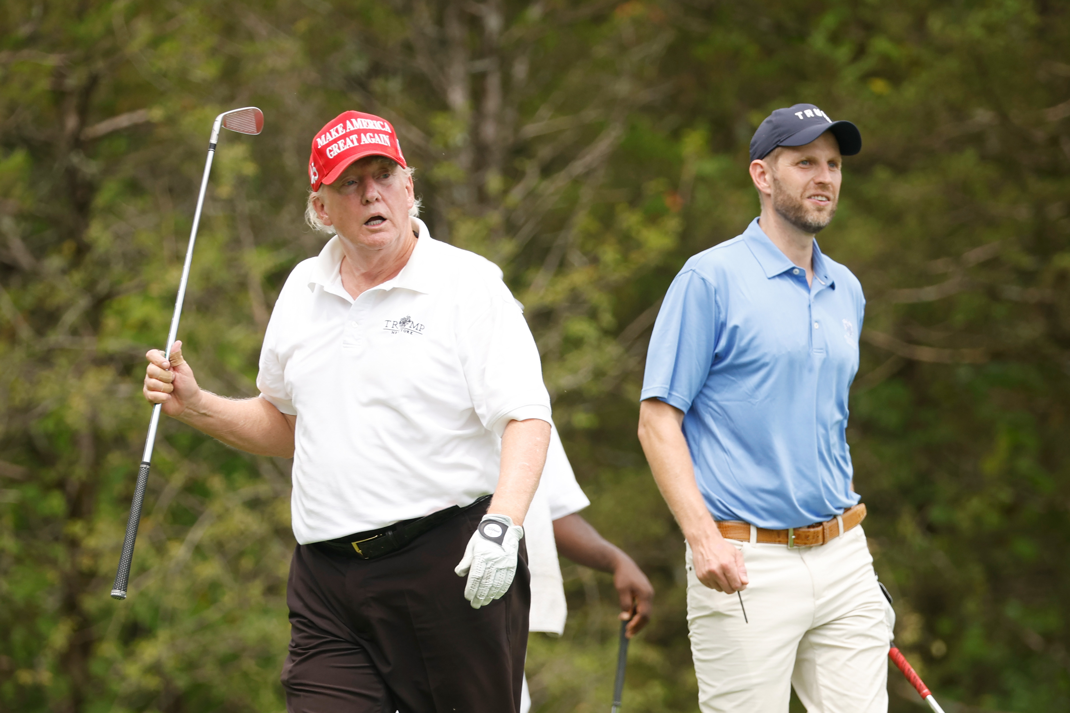 Donald Trump marche aux côtés de son fils, Eric Trump, pendant le tournoi, tenant un club. Son visage présente des rougeurs visibles autour des joues et du nez, tandis que le reste de sa peau semble plus clair.