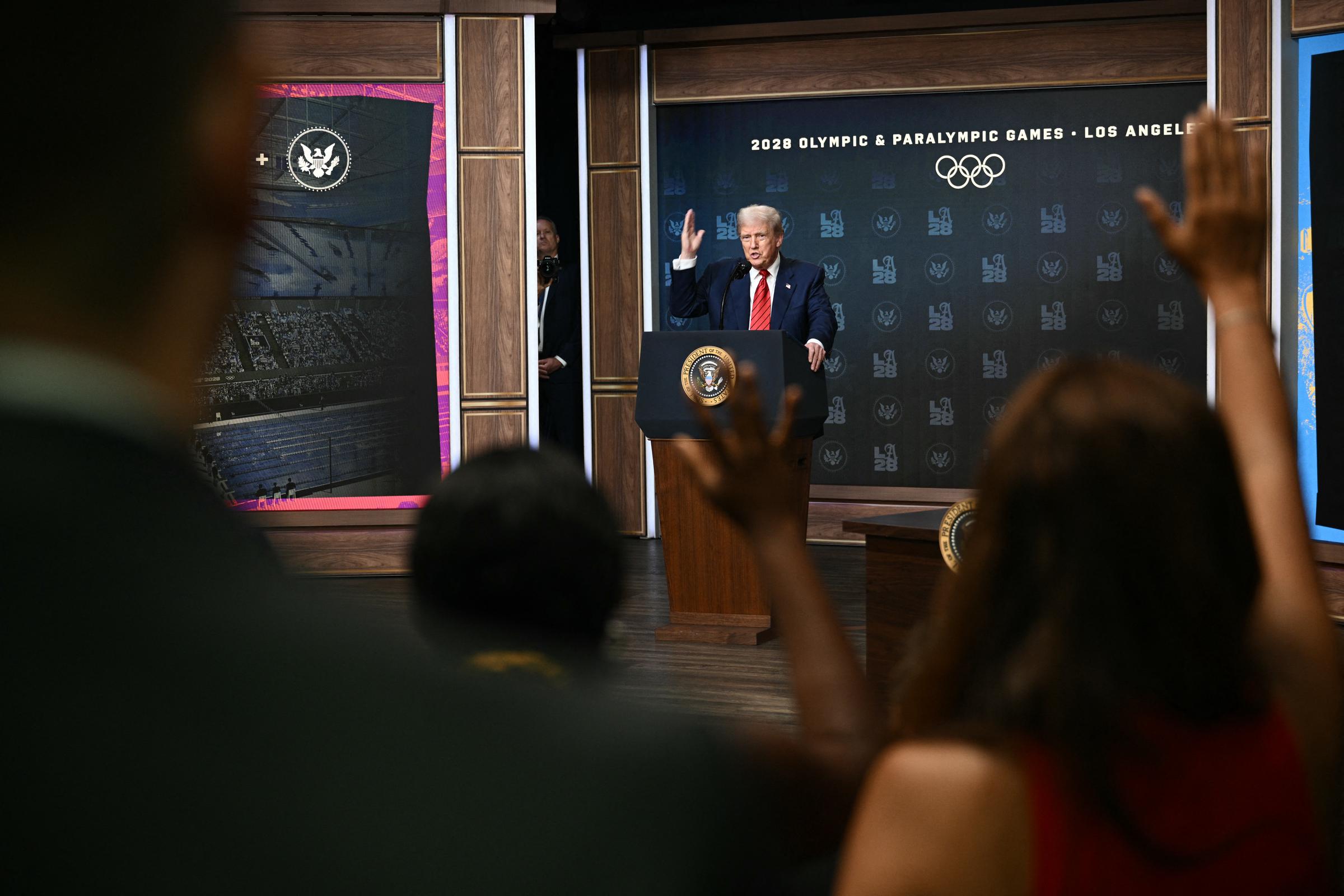 Après sa promenade sur le toit de la Maison Blanche, Donald Trump s'exprime depuis le podium de l'auditorium de South Court, répondant aux questions après avoir signé un décret visant à former un groupe de travail sur les Jeux olympiques de 2028, tandis que les membres de la presse lèvent la main.