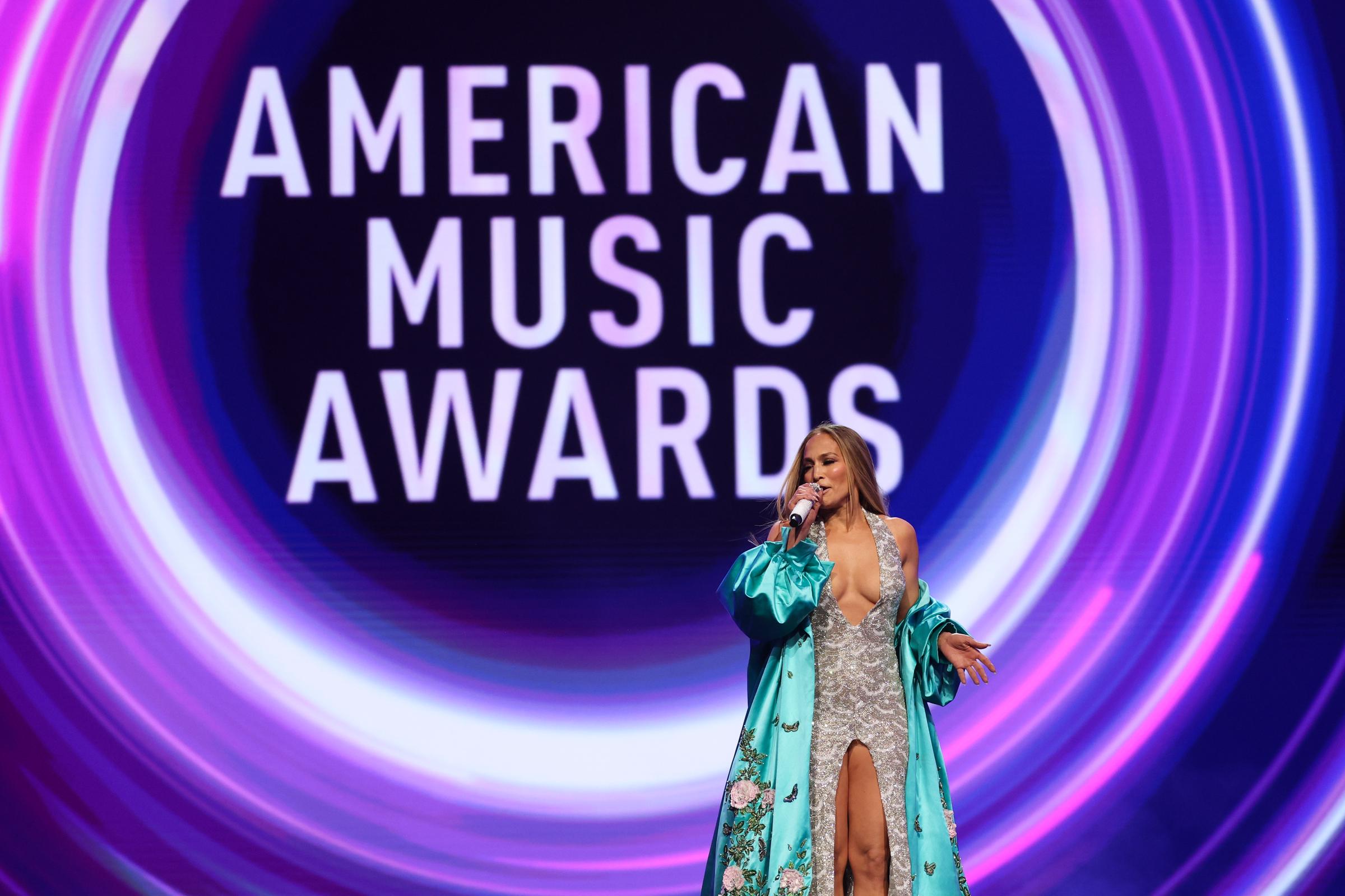 Au milieu de la présentation, Jennifer Lopez lâche une main tout en parlant, les paillettes de sa robe et la fluidité de sa robe brodée se détachant sur les graphiques audacieux et tourbillonnants de la scène des AMAs.