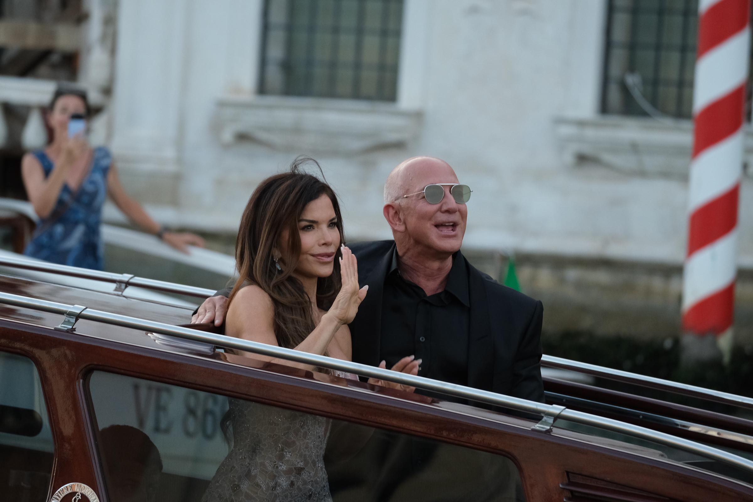 Lauren Sánchez lève la main dans un geste gracieux tandis que Jeff Bezos sourit à ses côtés. Tous deux se tiennent debout sur le bateau, tandis qu'une femme à l'arrière-plan immortalise l'instant avec son téléphone près d'un poteau rayé rouge et blanc.