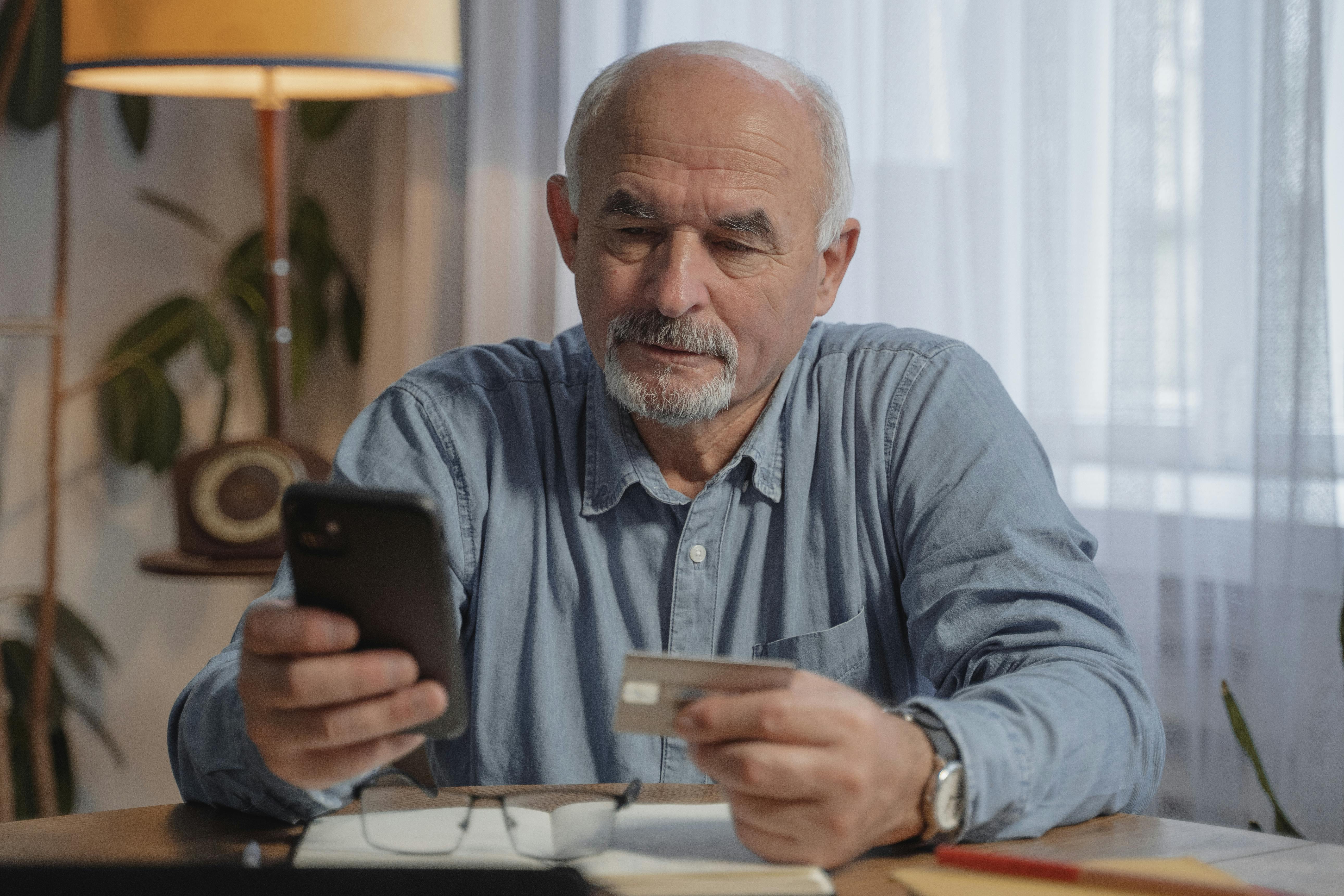 Un homme âgé tenant son téléphone dans une main et une carte bancaire dans l'autre | Source : Pexels