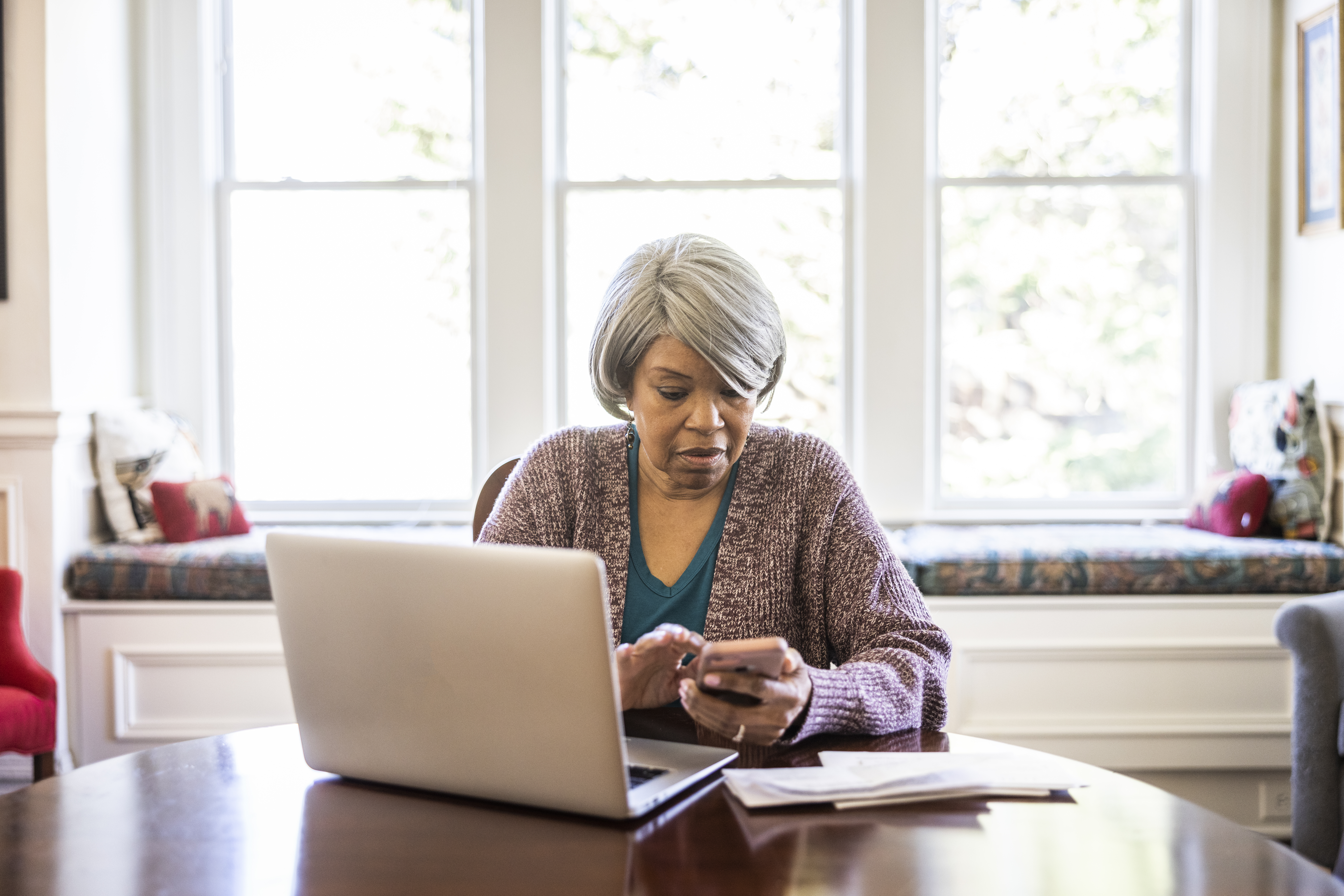 Femme âgée payant ses factures avec son ordinateur portable et son smartphone à la maison | Source : Getty Images