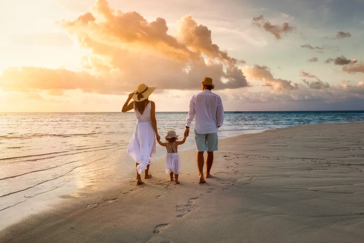 Une famille élégante en vêtements d'été blancs marchant main dans la main sur une plage paradisiaque tropicale pendant le coucher du soleil | Source : Shutterstock