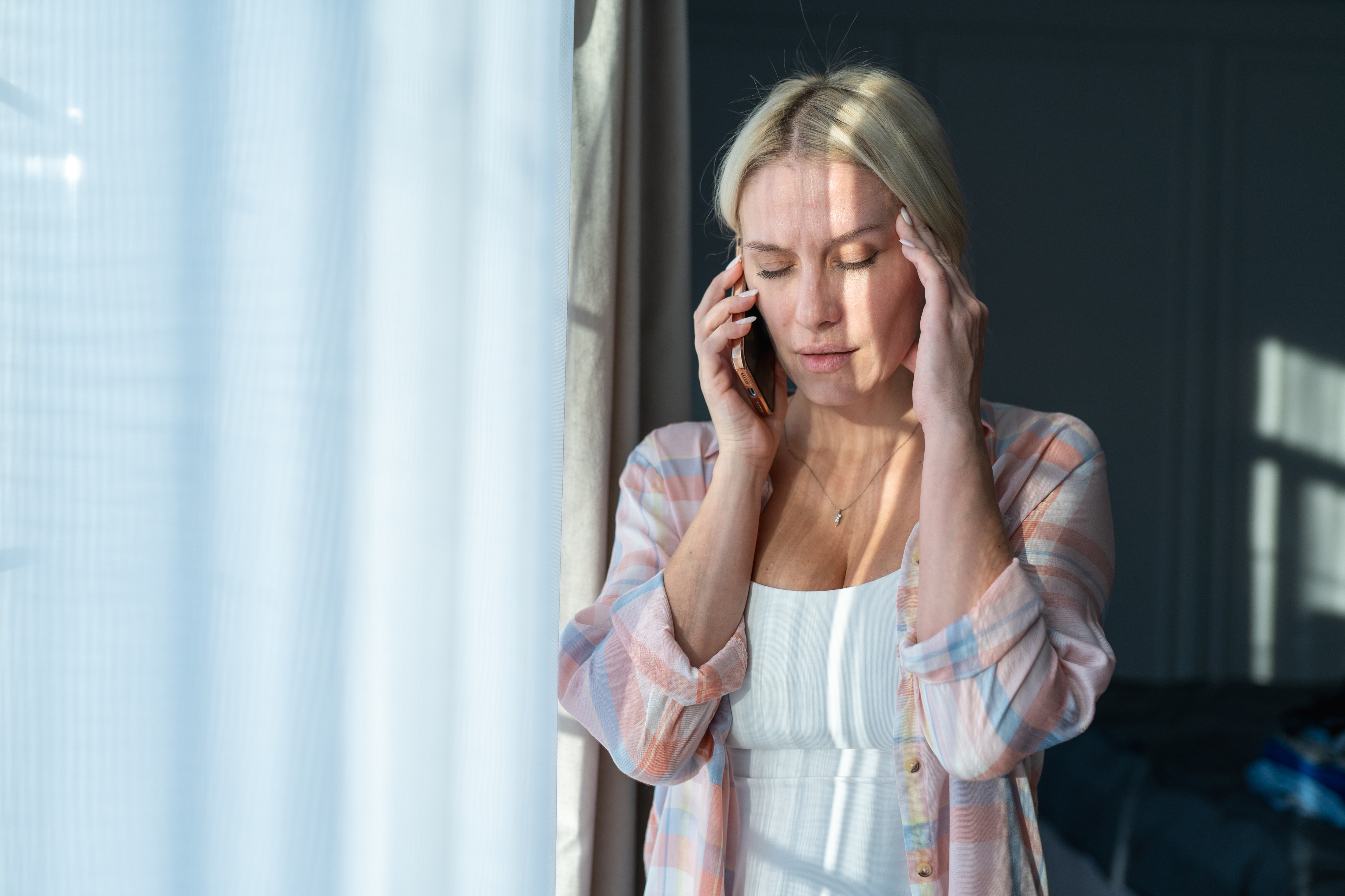 Maux de tête et désespoir. Une femme blonde est au téléphone, tout en se touchant la tête près d'une fenêtre lumineuse | Source : Getty Images