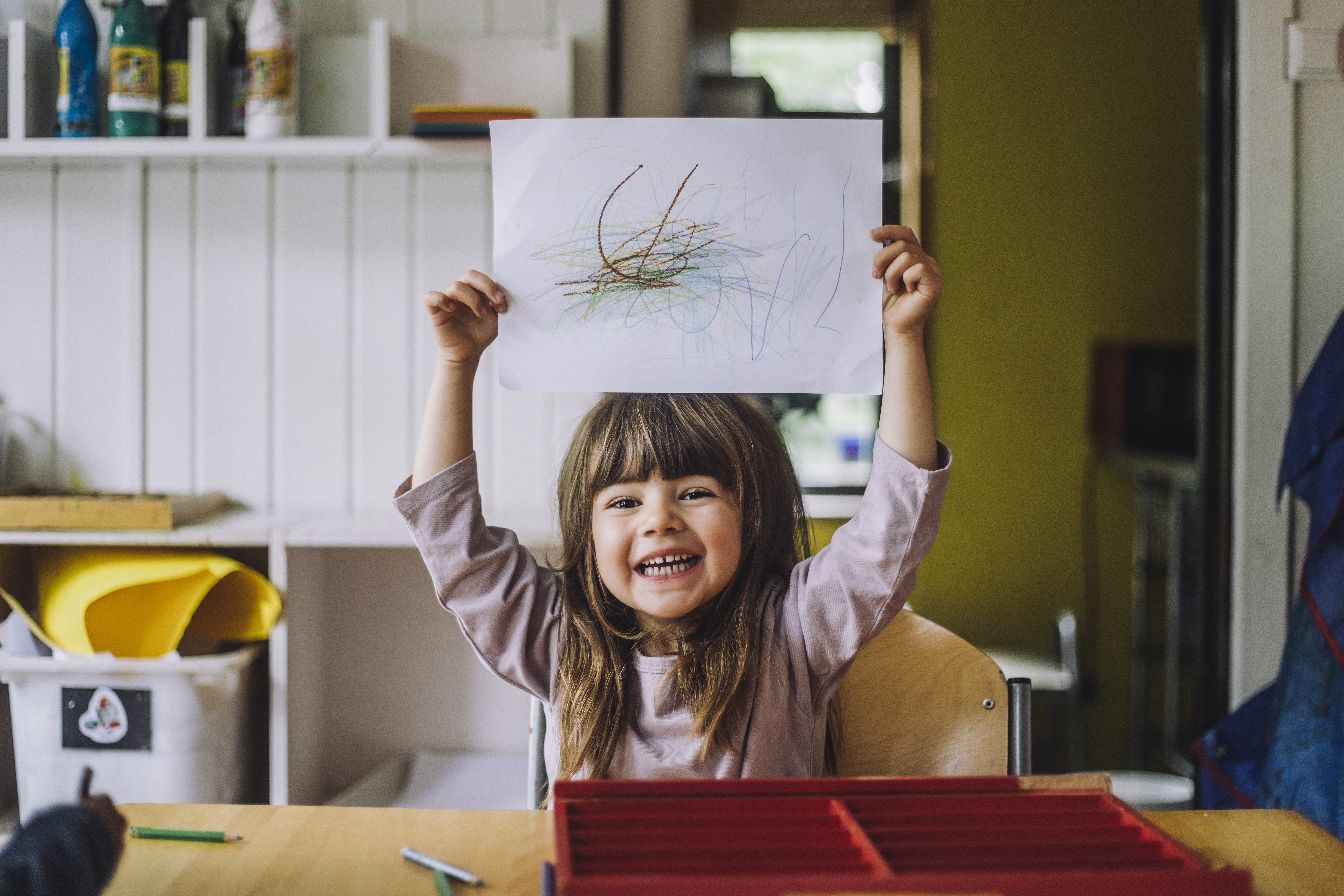 Petite fille heureuse montrant son gribouillage au crayon de couleur sur une feuille de papier à la maternelle | Source : Getty Images