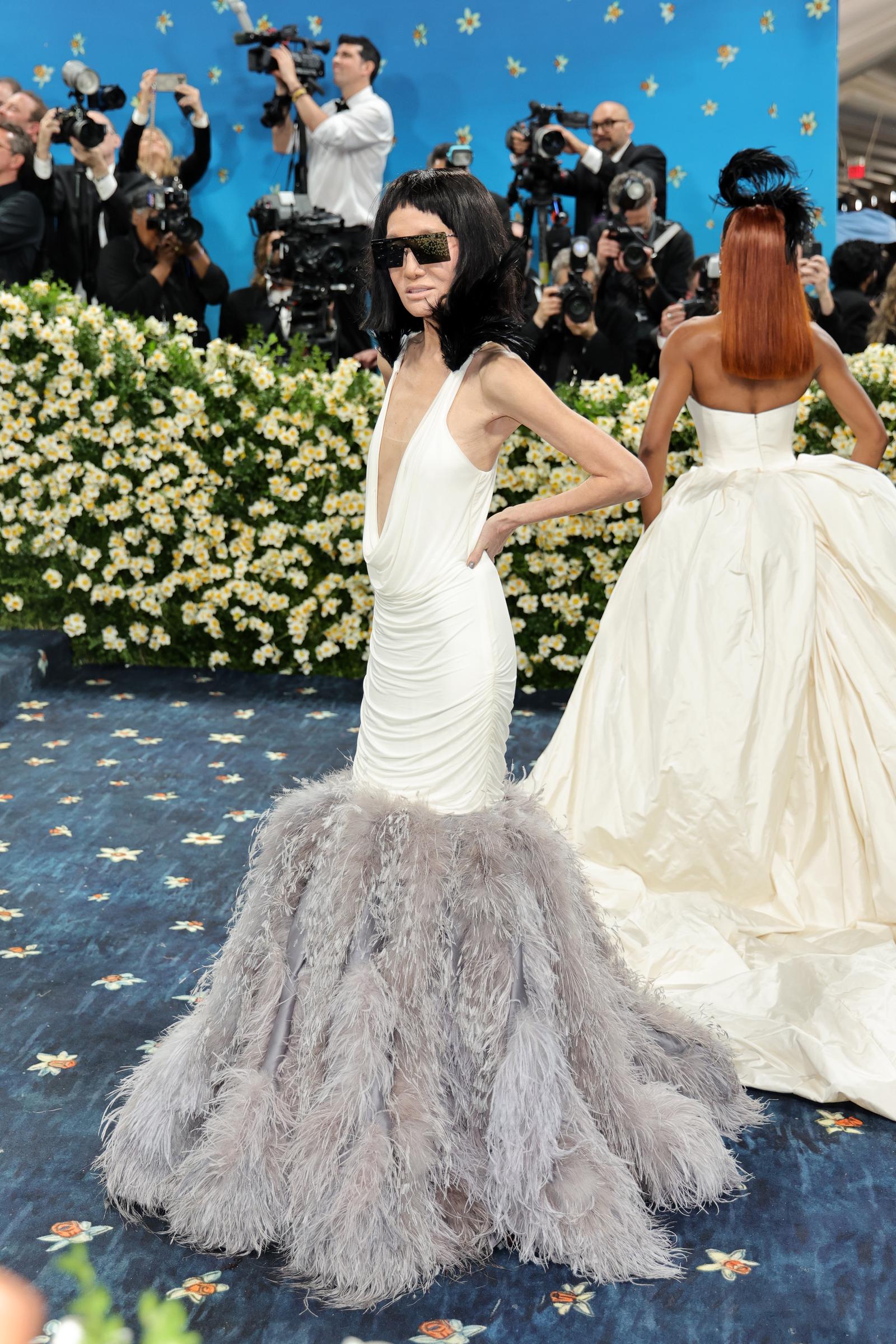 Avec des caméras qui clignotent derrière elle, Vera Wang pose de profil, mettant en valeur le drapé sculpté de la robe et la jupe volumineuse à plumes sur une toile de fond florale.