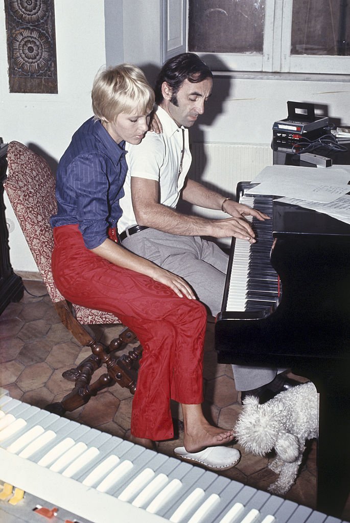 L'auteur-compositeur-interprète français d'origine arménienne Charles Aznavour joue un thème au piano ; à côté de lui est assise sa troisième épouse, la Suédoise Ulla Thorsell, qui caresse du pied un caniche blanc, tandis qu'en gros plan se détache un clavier. Italie, 1968. | Photo : Getty Images