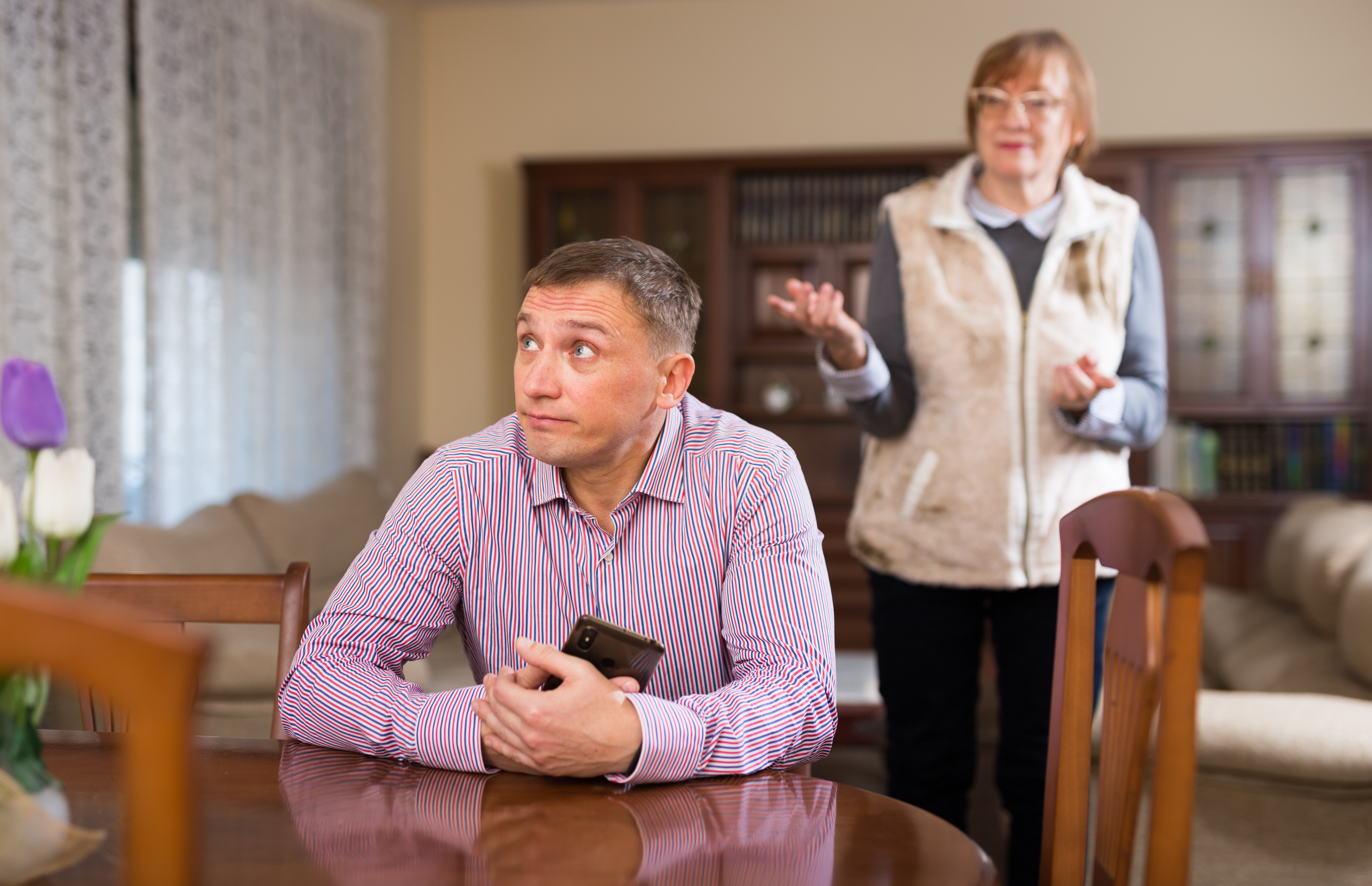 Un jeune homme tenant son téléphone et détournant le regard alors qu'il est assis à une table et qu'une dame plus âgée tente de lui parler en arrière-plan | Source : Shutterstock