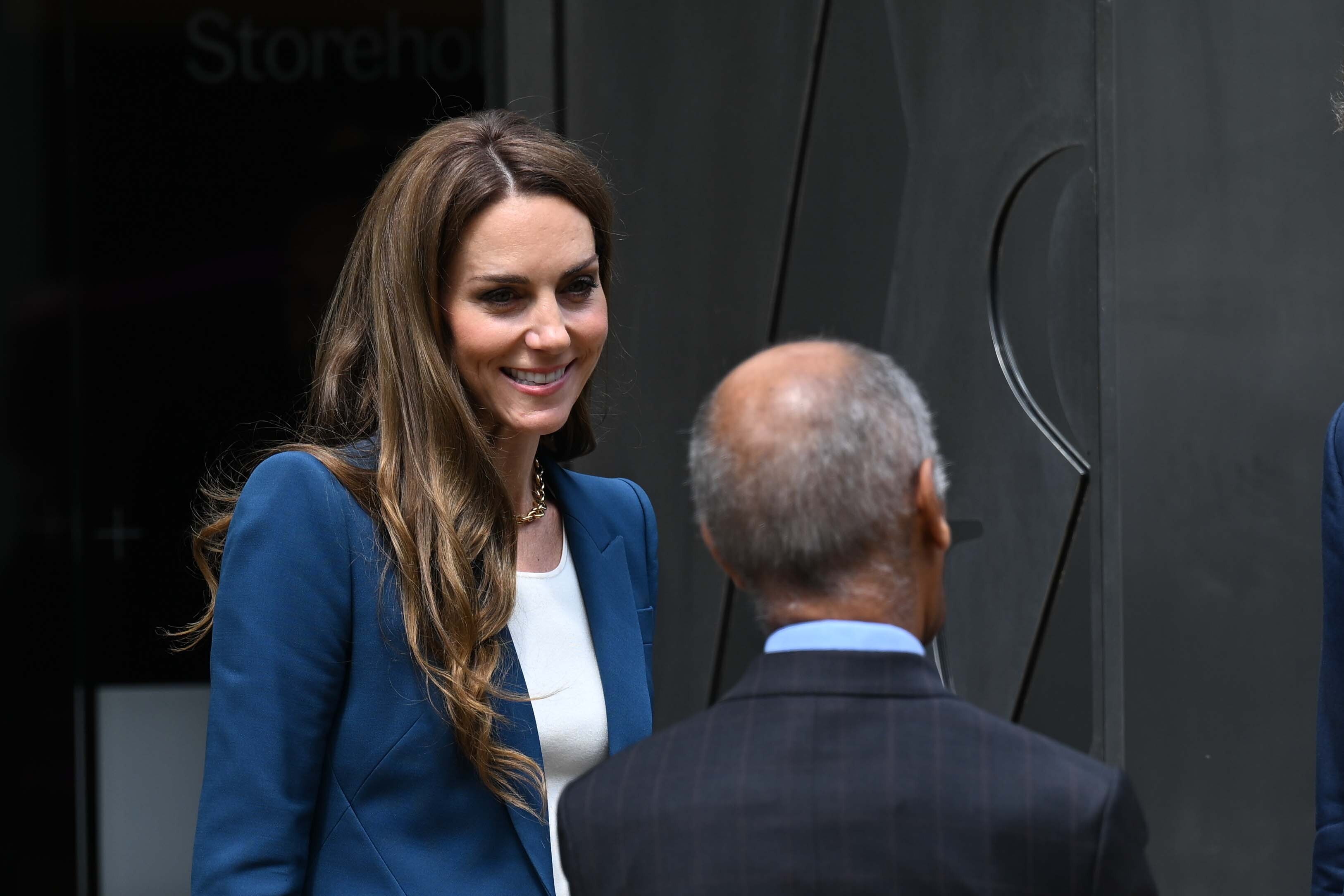 La princesse Catherine affiche un sourire subtil en écoutant attentivement, la tête légèrement inclinée dans une posture active et réceptive. Son expression lumineuse et son regard doux confèrent un air pensif à ce moment candide.