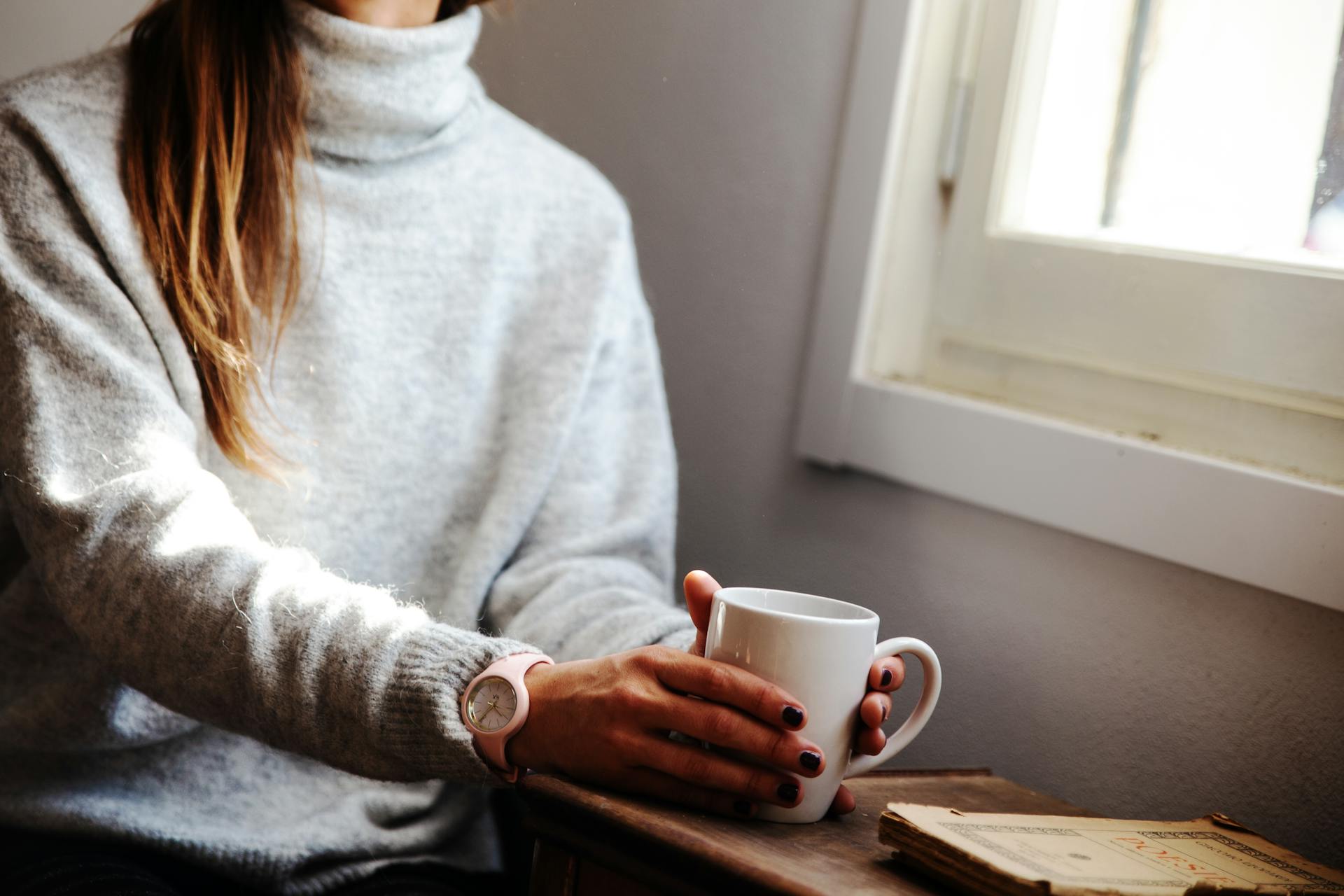 Une femme tenant une tasse de café et assise à côté de la fenêtre | Source : Pexels