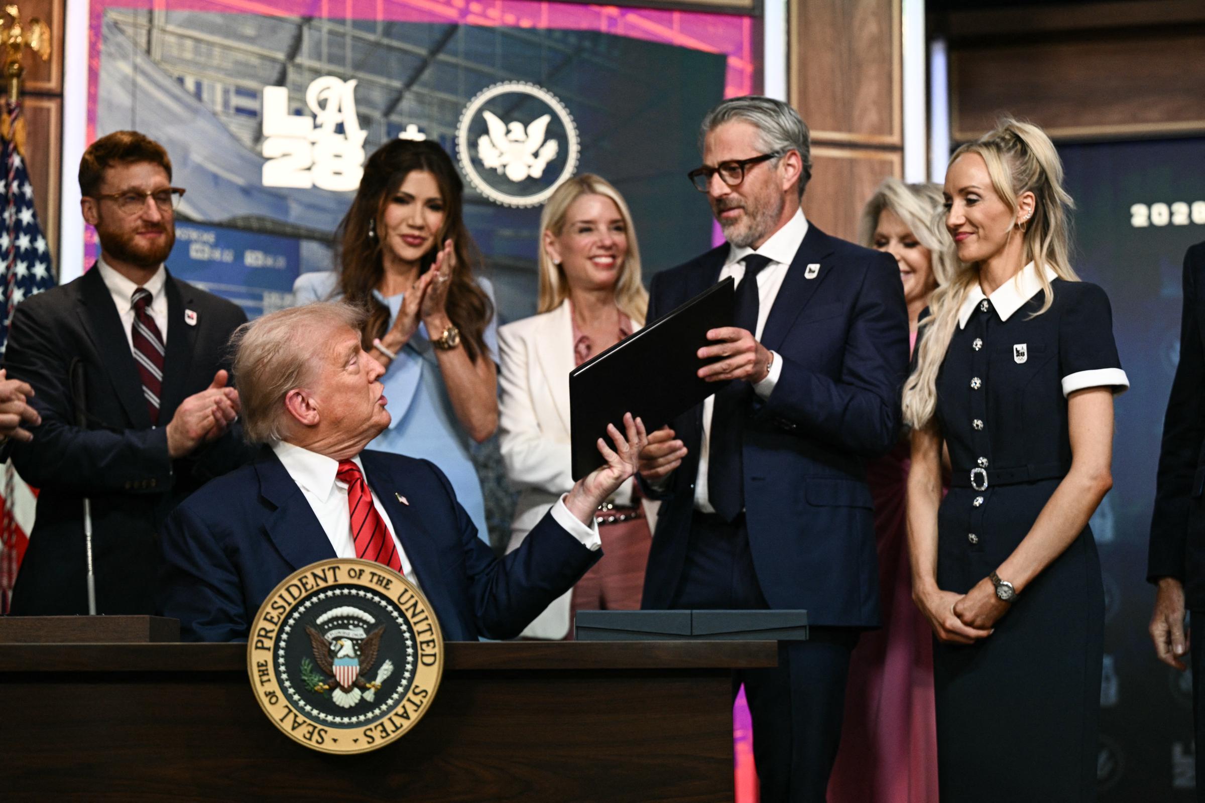 Donald Trump semble parler alors qu'il remet au président de LA 2028, Casey Wasserman, une copie du décret signé, sous le regard des participants qui sourient et applaudissent.
