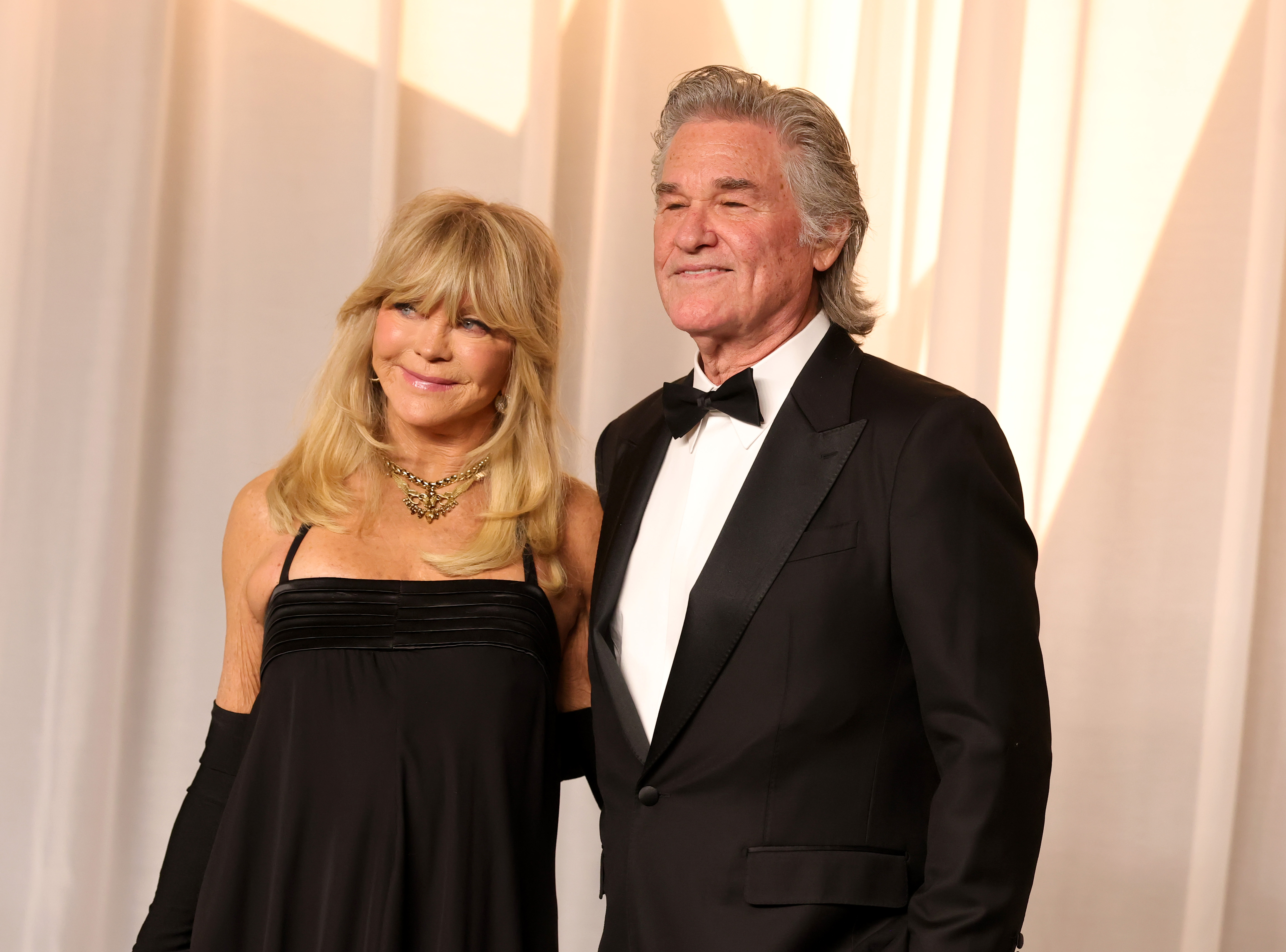 Hawn et Russell posent ensemble dans d'élégantes tenues de soirée : la robe à bretelles de Hawn, ses longs gants, ses bijoux imposants et sa chevelure blonde volumineuse contrastent avec le smoking sur mesure de Russell, sa chemise blanche impeccable et ses cheveux gris lissés en arrière.