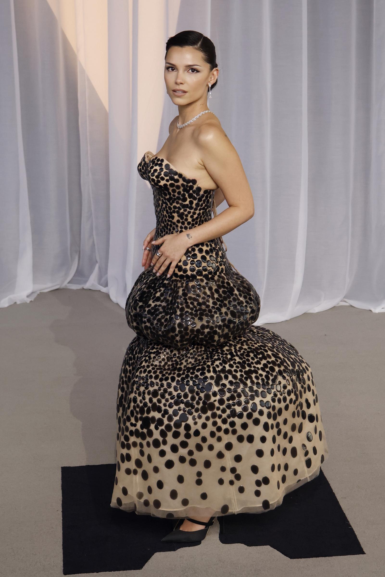 Inde Navarrette nous offre un look spectaculaire et avant-gardiste dans une robe sculptée sans bretelles, ornée de gros pois brillants sur un fond neutre. La silhouette volumineuse s'évase audacieusement au niveau des hanches et de l'ourlet bouffant en cascade, qu'elle met en valeur avec une coiffure relevée épurée et soignée, un collier ras-du-cou en diamants étincelants et des escarpins noirs à bout pointu.