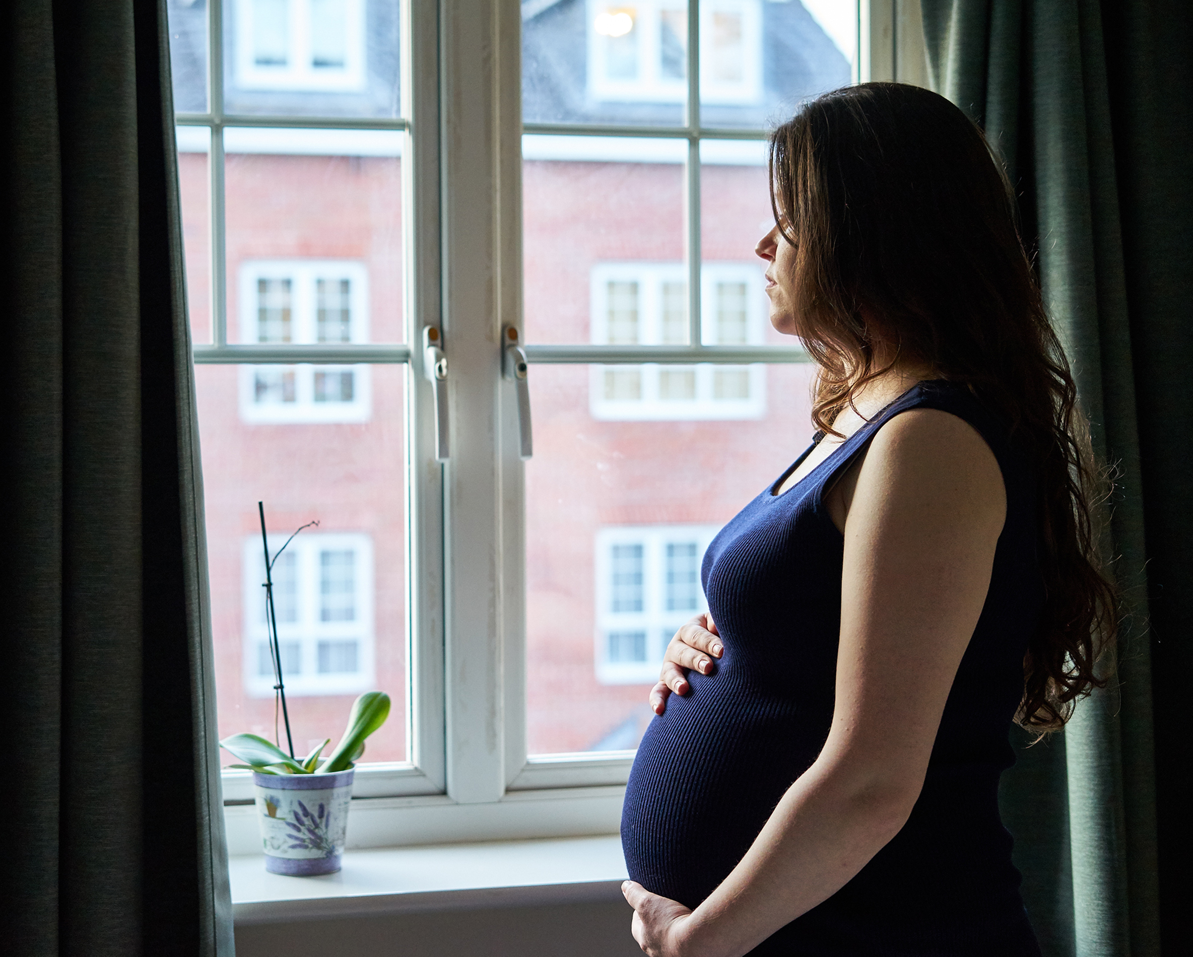 Une femme lourdement enceinte se tenant debout et tenant son ventre tout en regardant à l'extérieur d'une fenêtre | Source : Shutterstock