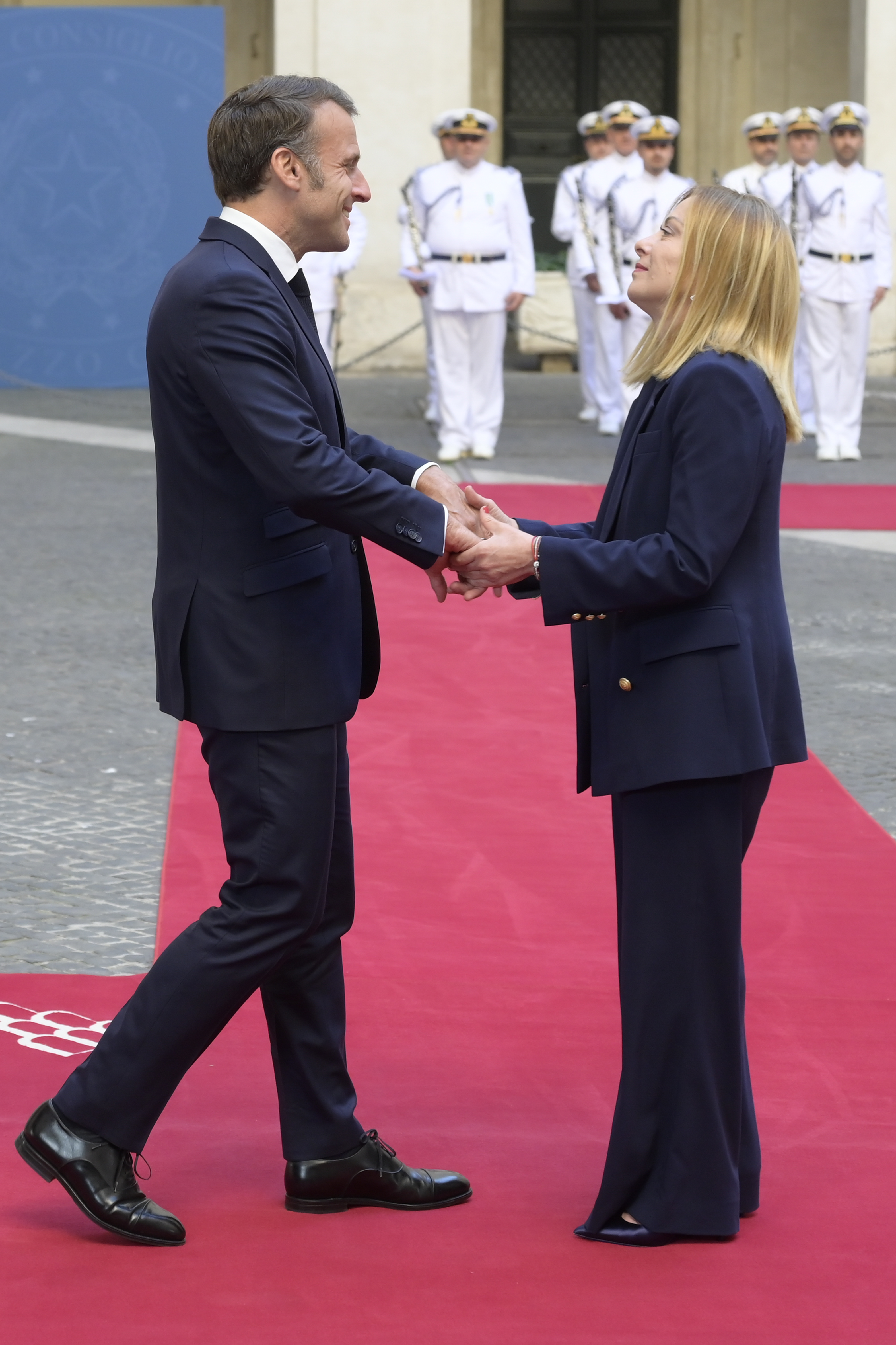 Emmanuel Macron et Giorgia Meloni se serrent fermement la main alors qu'ils se tiennent debout sur un tapis rouge devant le Palazzo Chigi. Macron affiche un large sourire tandis que Giorgia lève les yeux vers lui, tous deux entourés de gardes d'honneur en uniformes blancs.