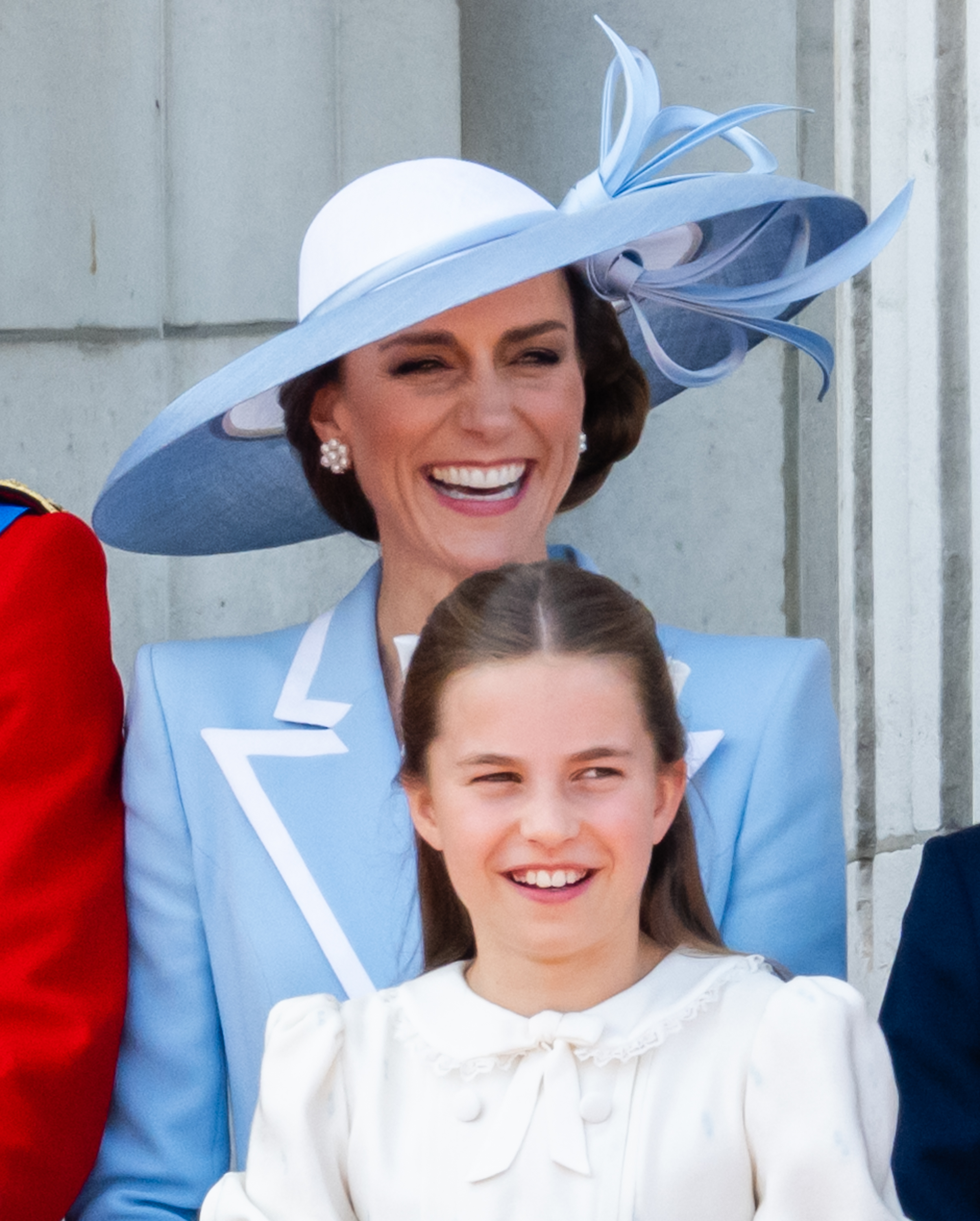 La princesse Charlotte de Galles et Catherine, princesse de Galles, rient joyeusement sur le balcon du palais de Buckingham en regardant le défilé aérien de la RAF lors de la cérémonie du « Trooping the Colour », le 13 juin 2026, à Londres, en Angleterre. Charlotte arbore un large sourire dans une robe ivoire ornée d’un col en dentelle à volants et d’un nœud à l’encolure, debout devant Catherine, qui porte une robe-manteau bleu pâle sur mesure rehaussée de revers blancs impeccables, des boucles d’oreilles en grappes de perles et un chapeau architectural signé Philip Treacy, garni de boucles de ruban sculptées. | Source : Getty Images