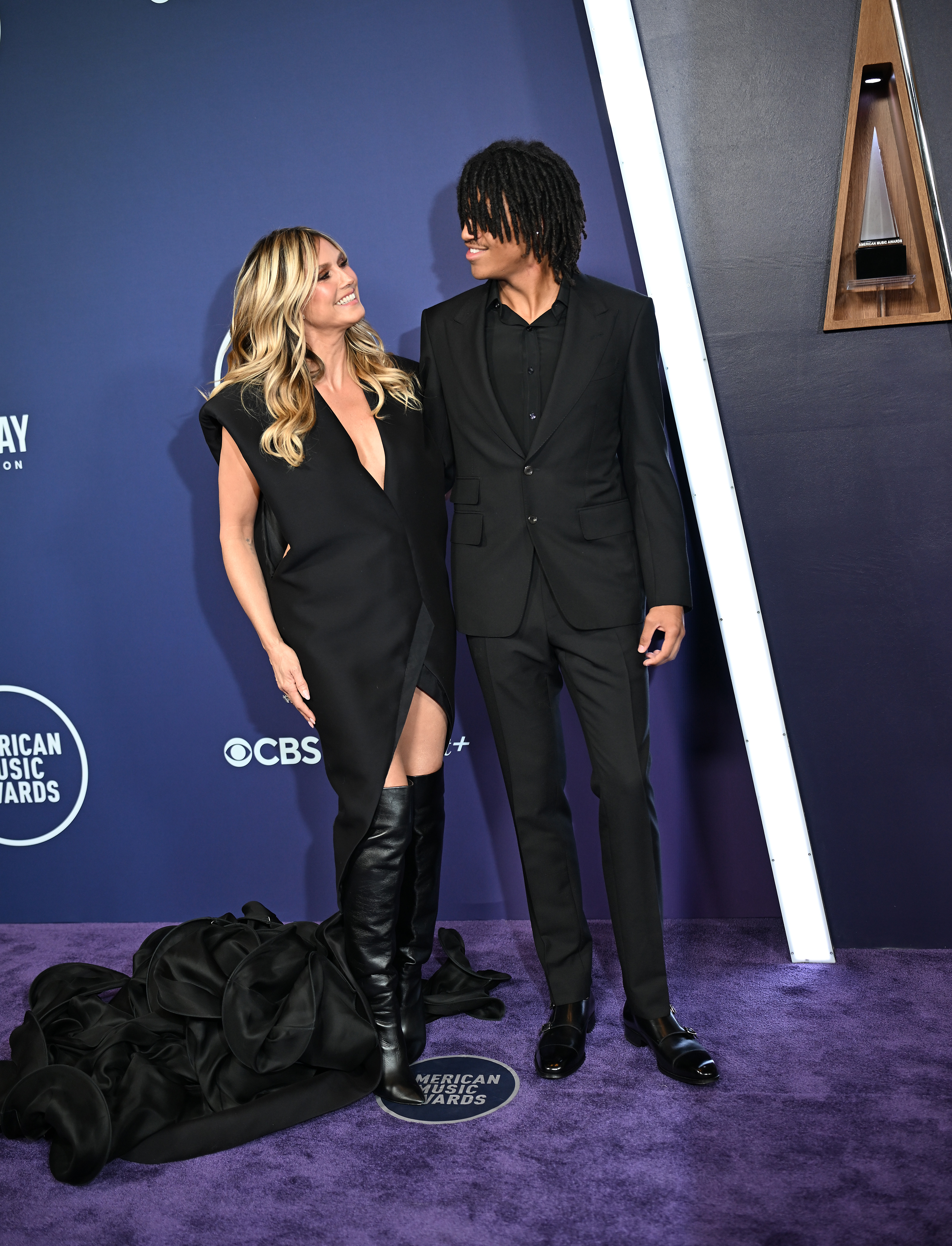 Heidi Klum partage un moment chaleureux avec son fils, Henry Samuel, alors qu'ils échangent des sourires sur le tapis rouge des AMAs - sa traîne dramatique se confond avec son costume noir bien coupé.