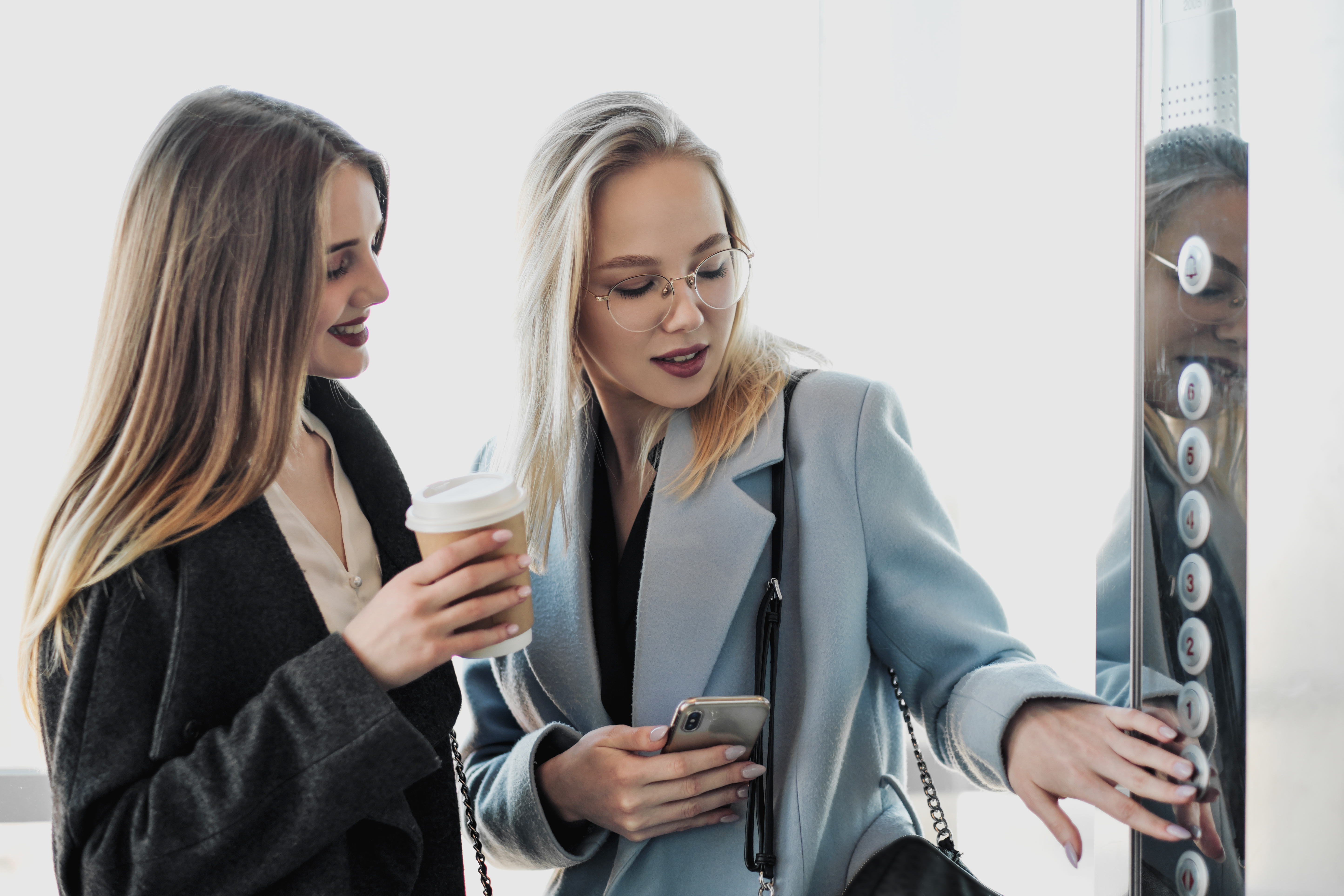 Dos chicas guapas, una rubia y una morena, llevan un abrigo en un ascensor de cristal. | Fuente: Shutterstock