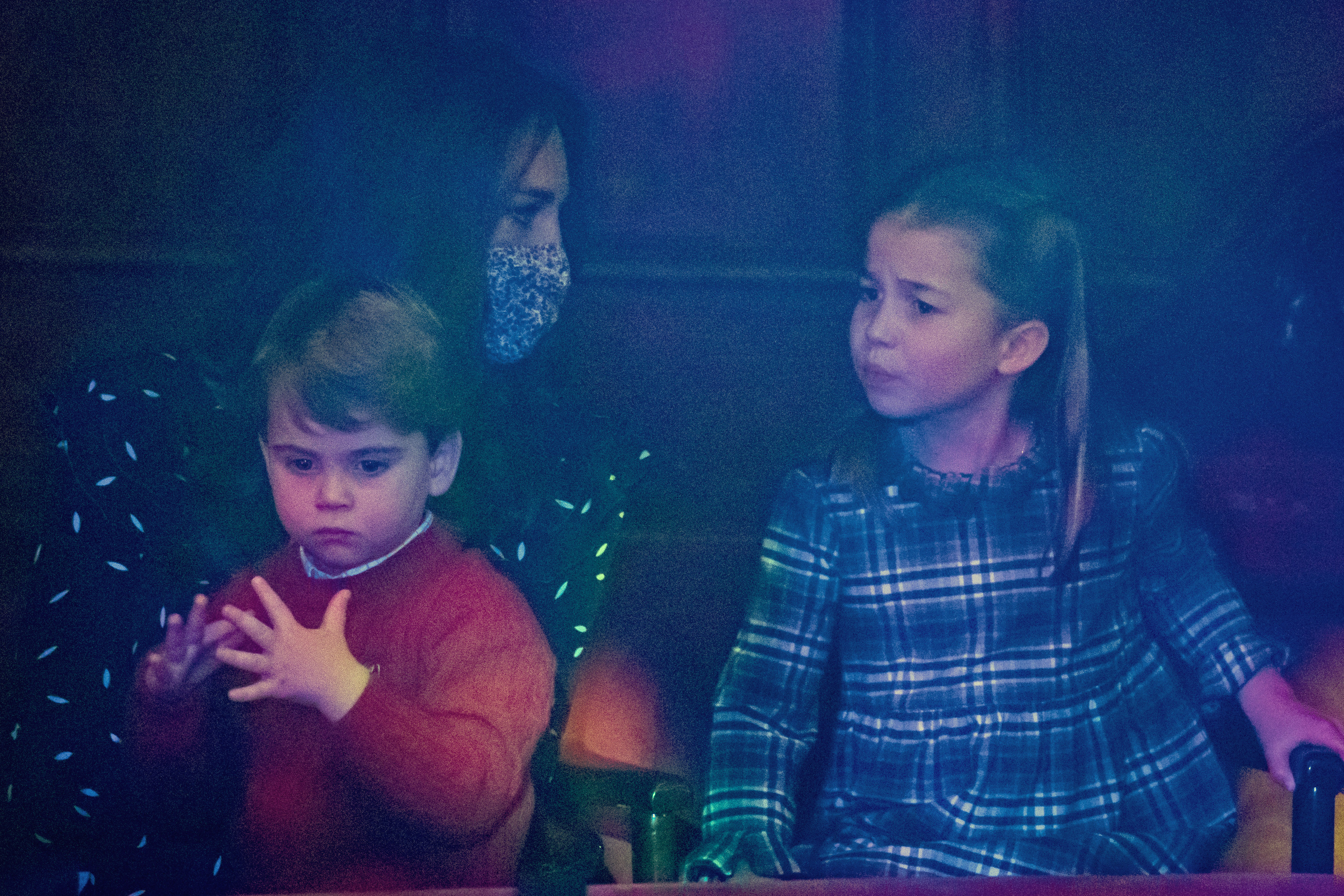 La princesse Charlotte et le prince Louis regardent depuis le public une représentation spéciale de pantomime au London Palladium Theatre, le 11 décembre 2020, à Londres, en Angleterre, organisée pour remercier les travailleurs essentiels et leurs familles pour leurs efforts pendant la pandémie. Charlotte est assise, toute attentive, dans une robe en tartan bleu et vert, à côté de Louis qui porte un pull rouge confortable à col rond par-dessus une chemise à col, tandis que leur mère Catherine, duchesse de Cambridge, les observe, vêtue d'une robe imprimée et d'un masque de protection. | Source : Getty Images