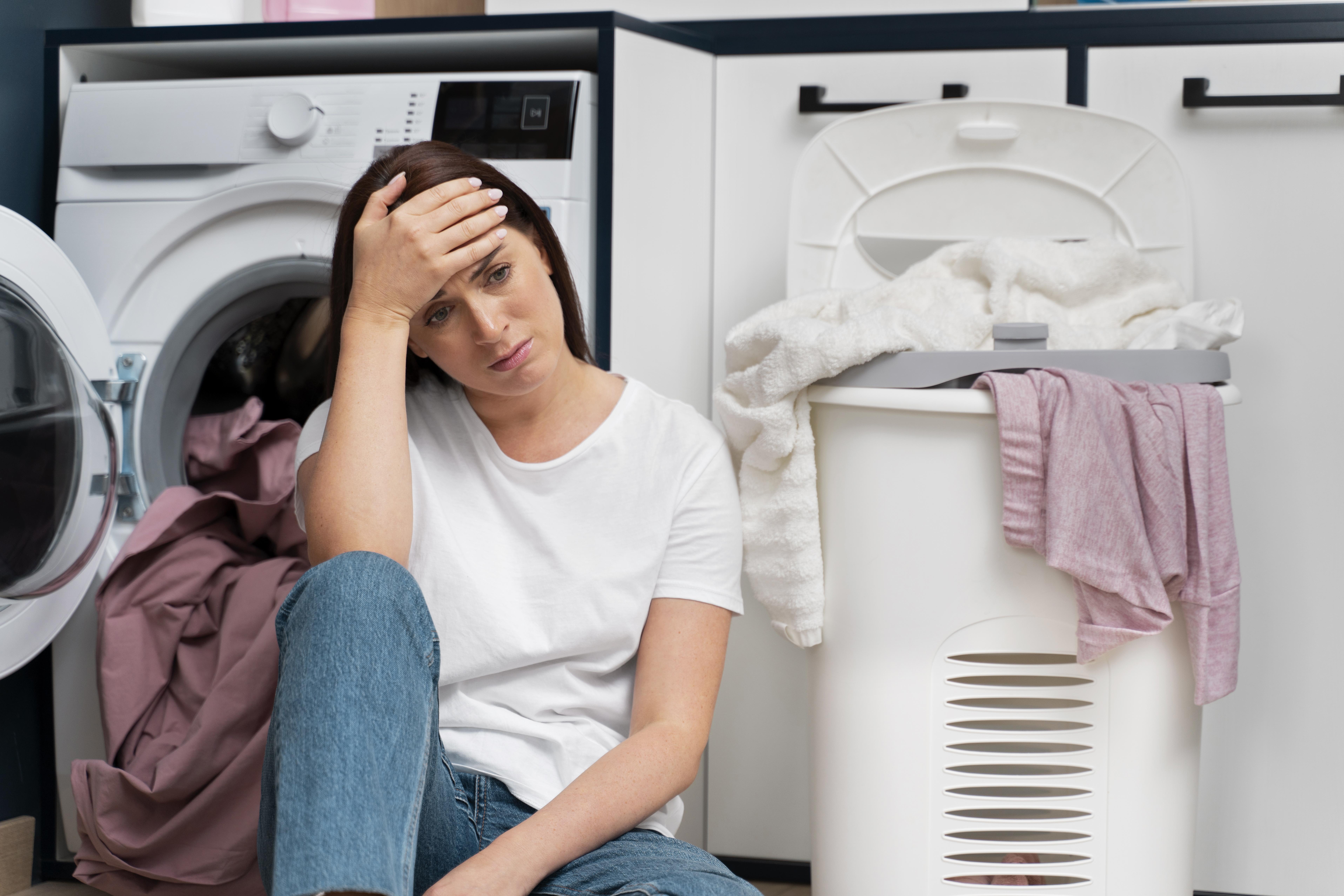 Une femme a l'air contrariée alors qu'elle s'appuie sur une machine à laver, entourée de linge | Source : Freepik