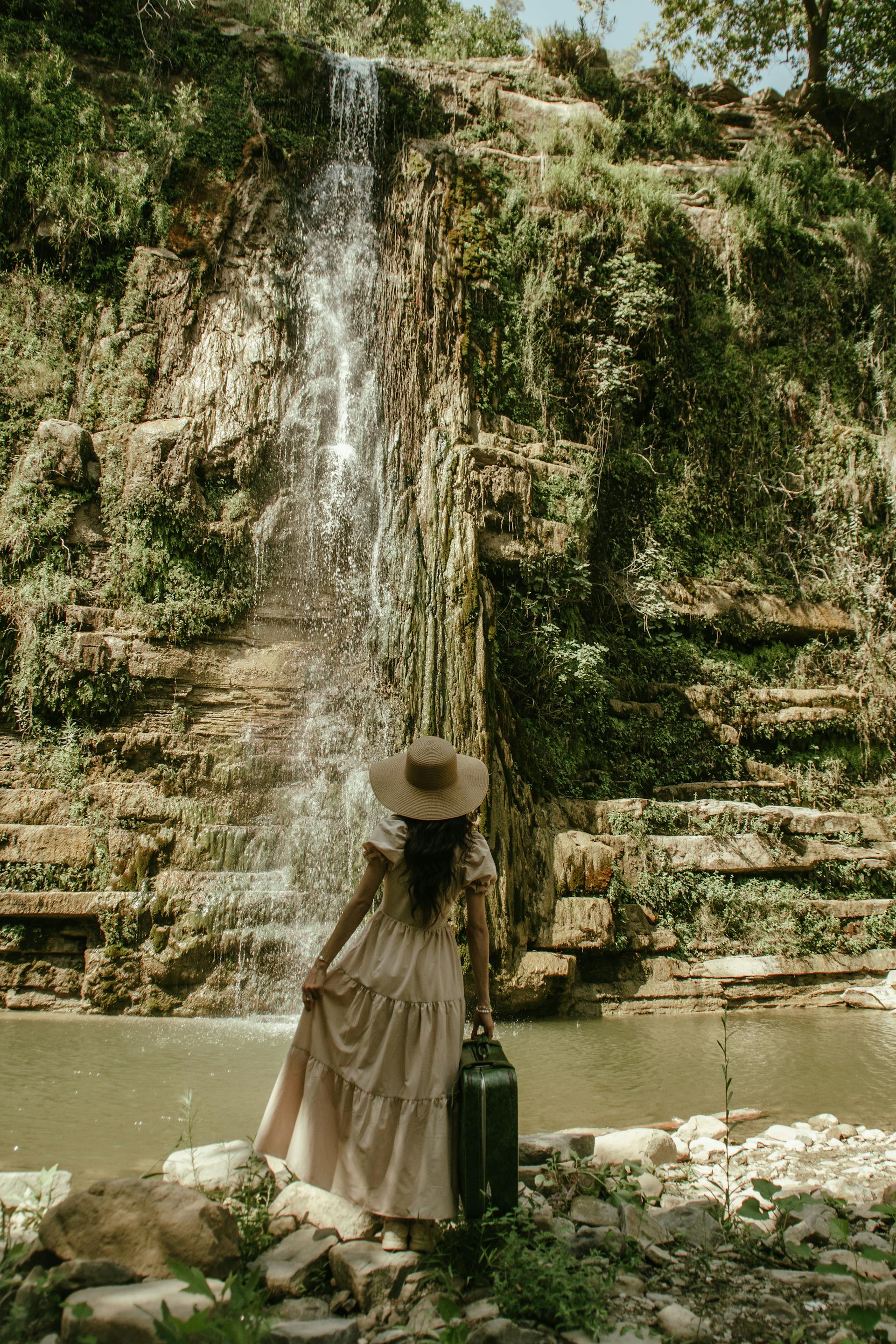 Une femme en robe beige et chapeau de paille qui regarde une cascade | Source : Pexels