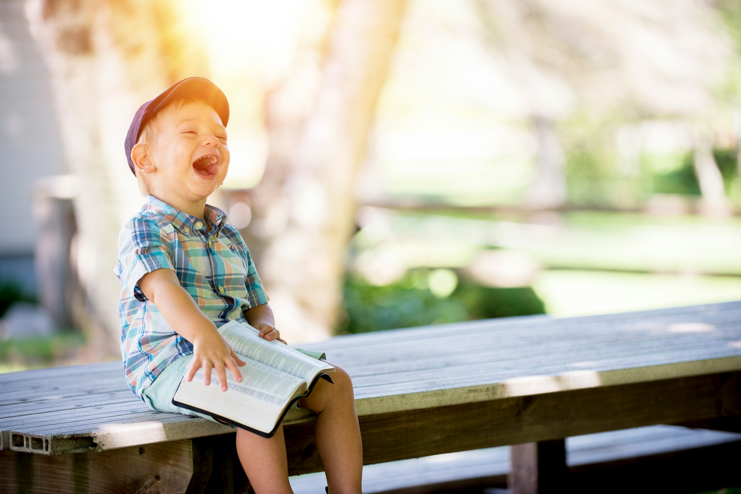 A little boy laughs while sitting on a bench and holding a book | Source: Unsplash