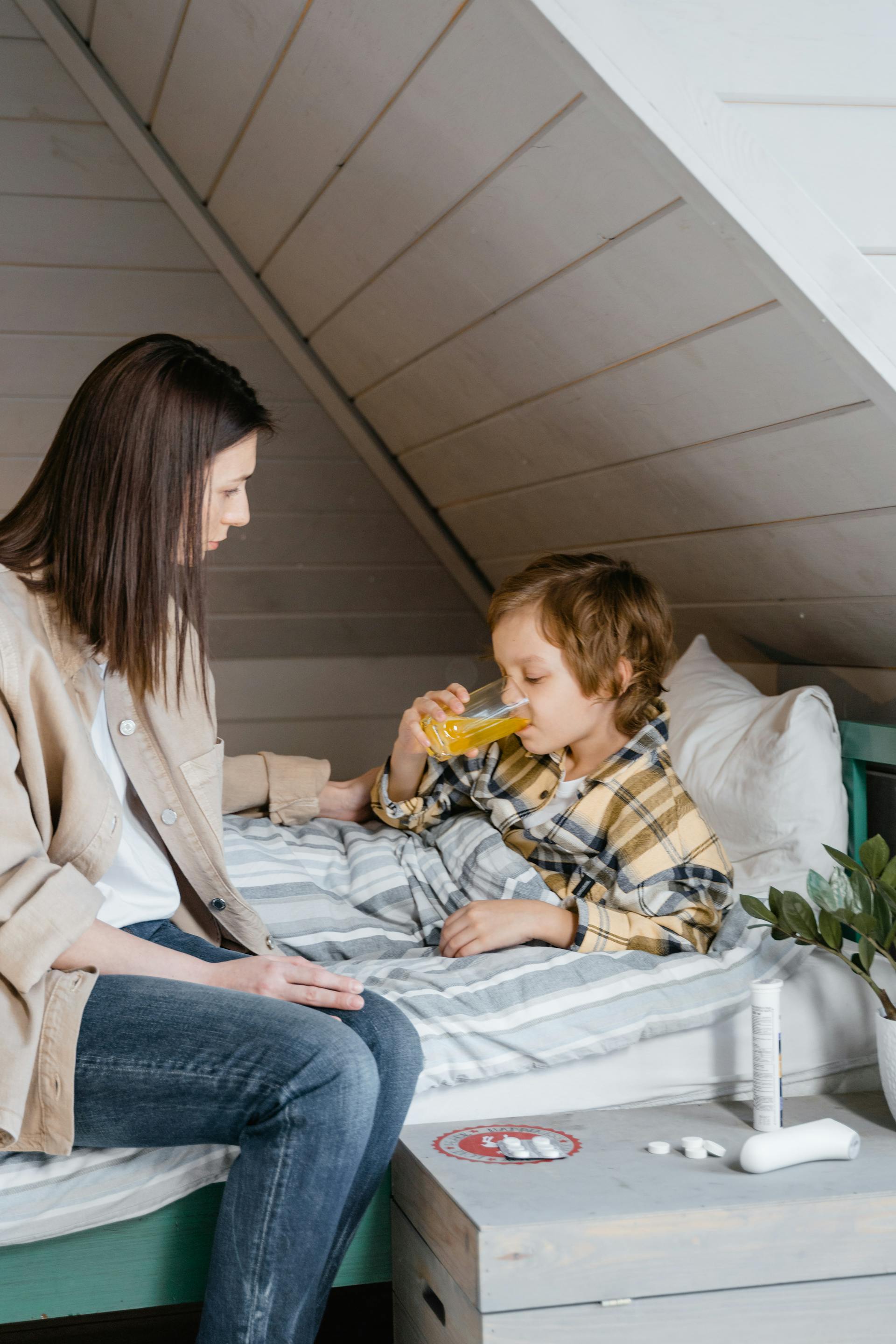 Une femme regarde son jeune fils qui boit un jus de fruit au lit | Source : Pexels