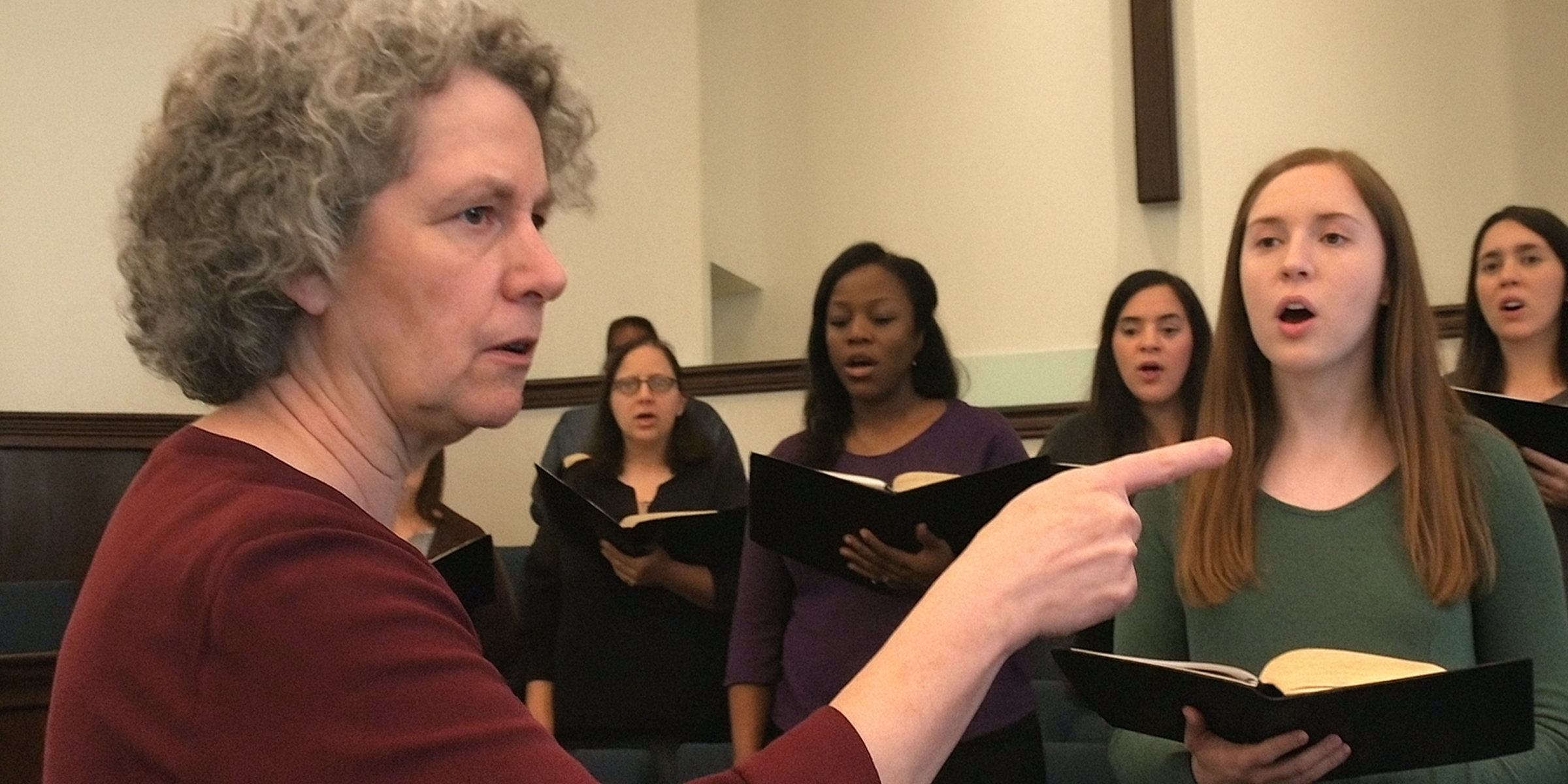 Une femme âgée et sévère interrompt la répétition d'une chorale en la pointant du doigt sous le regard de chanteurs stupéfaits | Source : The Celebritist