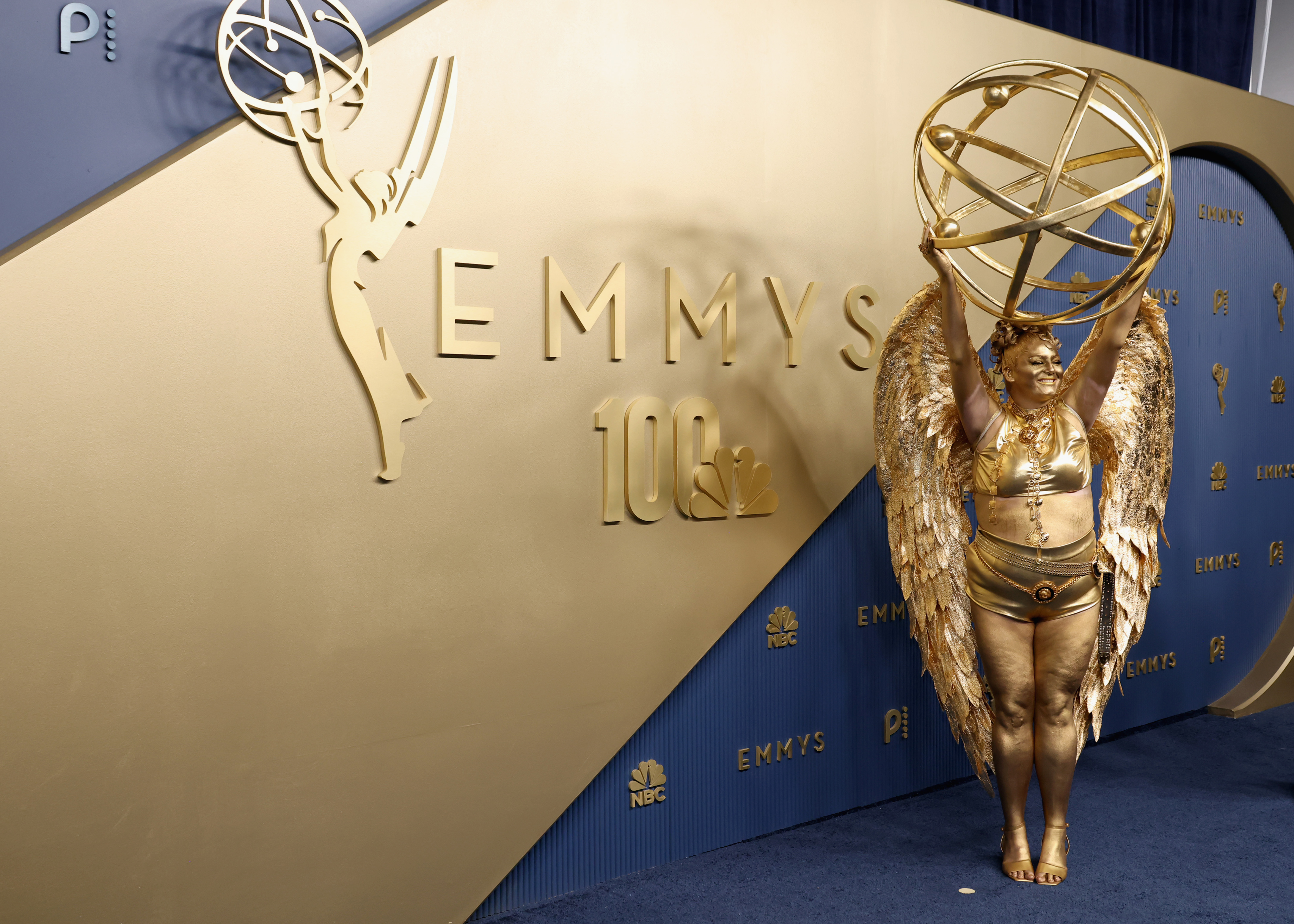 Meg Stalter pose dans sa tenue dorée métallisée et ses ailes sculptées devant le décor bleu et or des Emmy Awards, mettant en valeur sa coiffure sophistiquée et son maquillage doré sur le corps. | Source : Getty Images