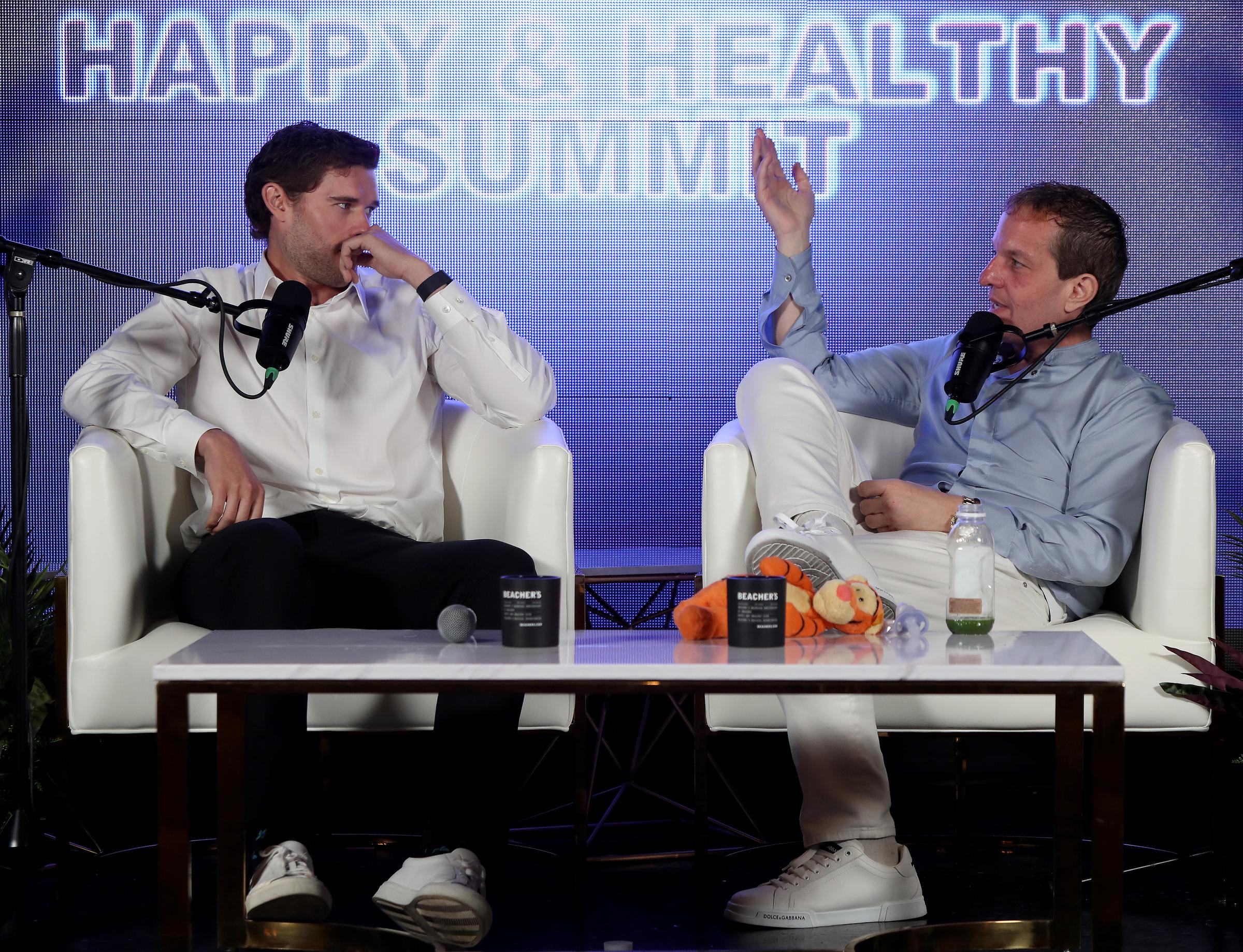 Christopher Schwarzenegger écoute attentivement les propos de Jeff Beacher pendant leur discussion. Les deux hommes sont assis l'un en face de l'autre sur la scène, en pleine discussion sur le développement personnel et la santé.