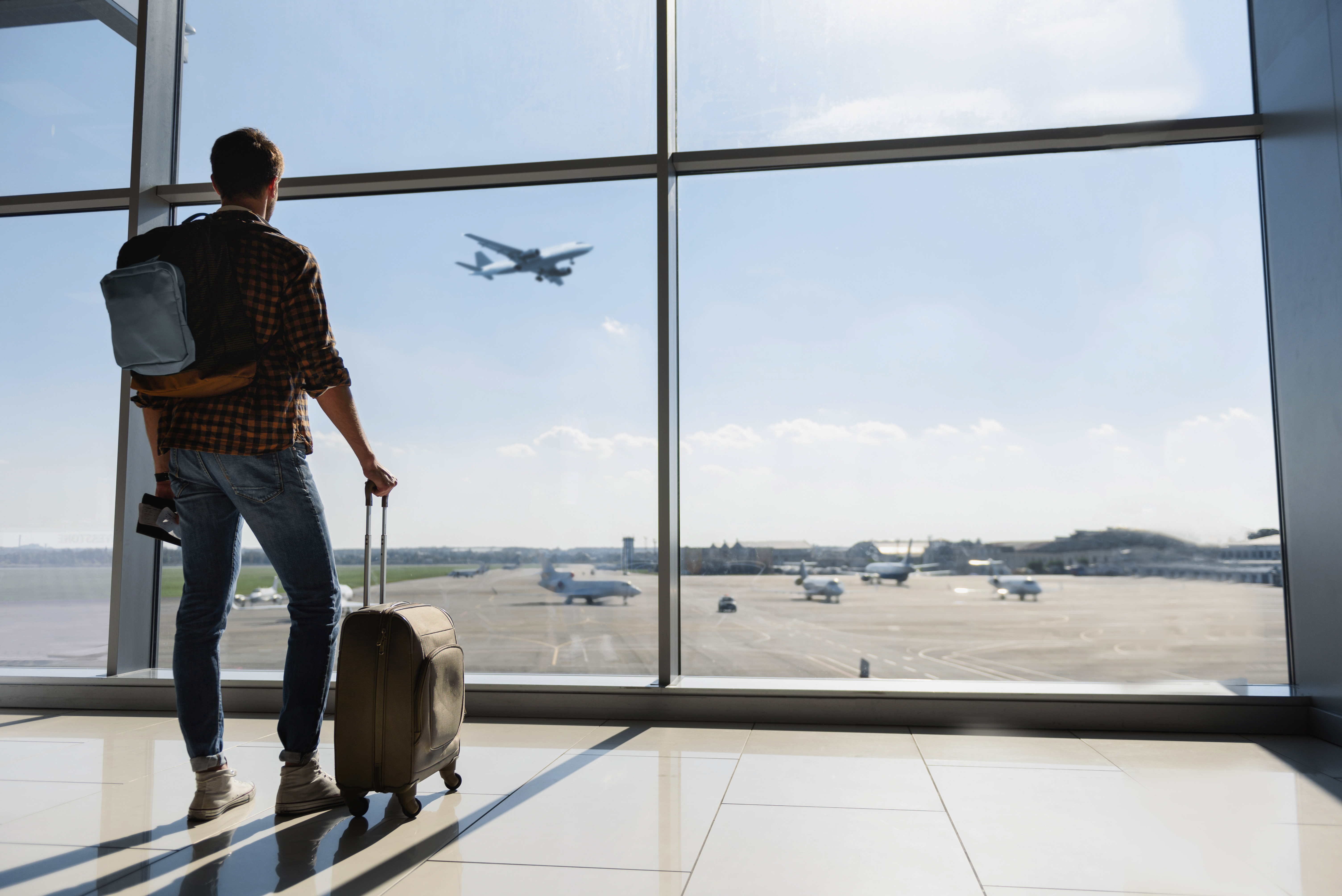 Un jeune homme avec ses bagages se tenant près d'une fenêtre d'aéroport et regardant un avion avant le départ | Source : Shutterstock