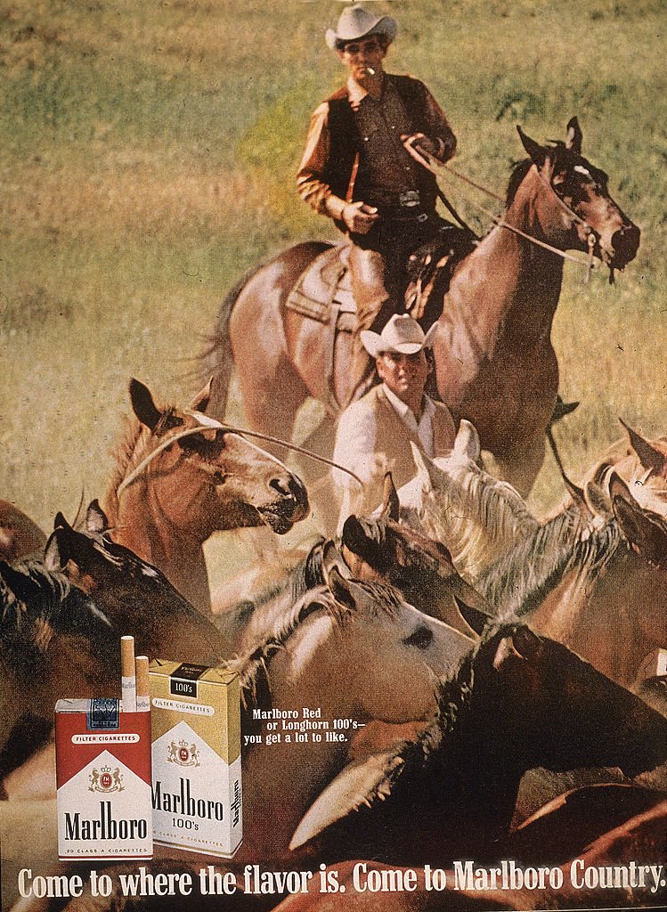 Image de deux cow-boys'Marlboro Man' rassemblant des chevaux dans une publicité pour des cigarettes Marlboro dans un magazine, années 1970| Source : Getty Images.