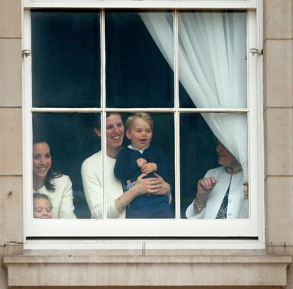 Le prince George de Cambridge retenu à une fenêtre du palais de Buckingham par sa nounou Maria Teresa Turrion Borrallo pour regarder Trooping the Colour le 13 juin 2015 à Londres, en Angleterre. La cérémonie est le défilé annuel d'anniversaire de la reine Elizabeth II. І Source : Getty Images