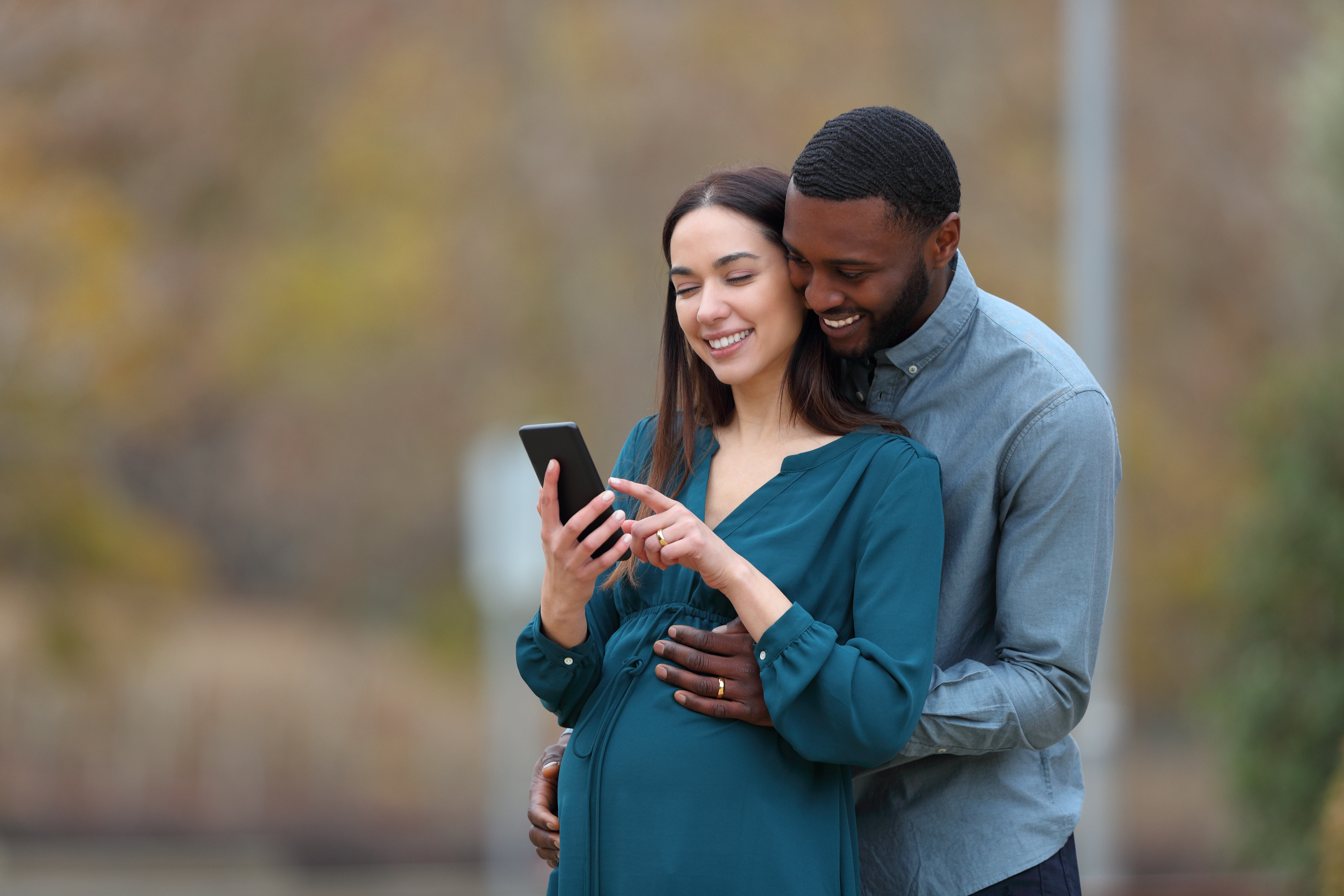 Un couple interracial dont la femme enceinte et son mari regardent leur smartphone dans un parc | Source : Shutterstock