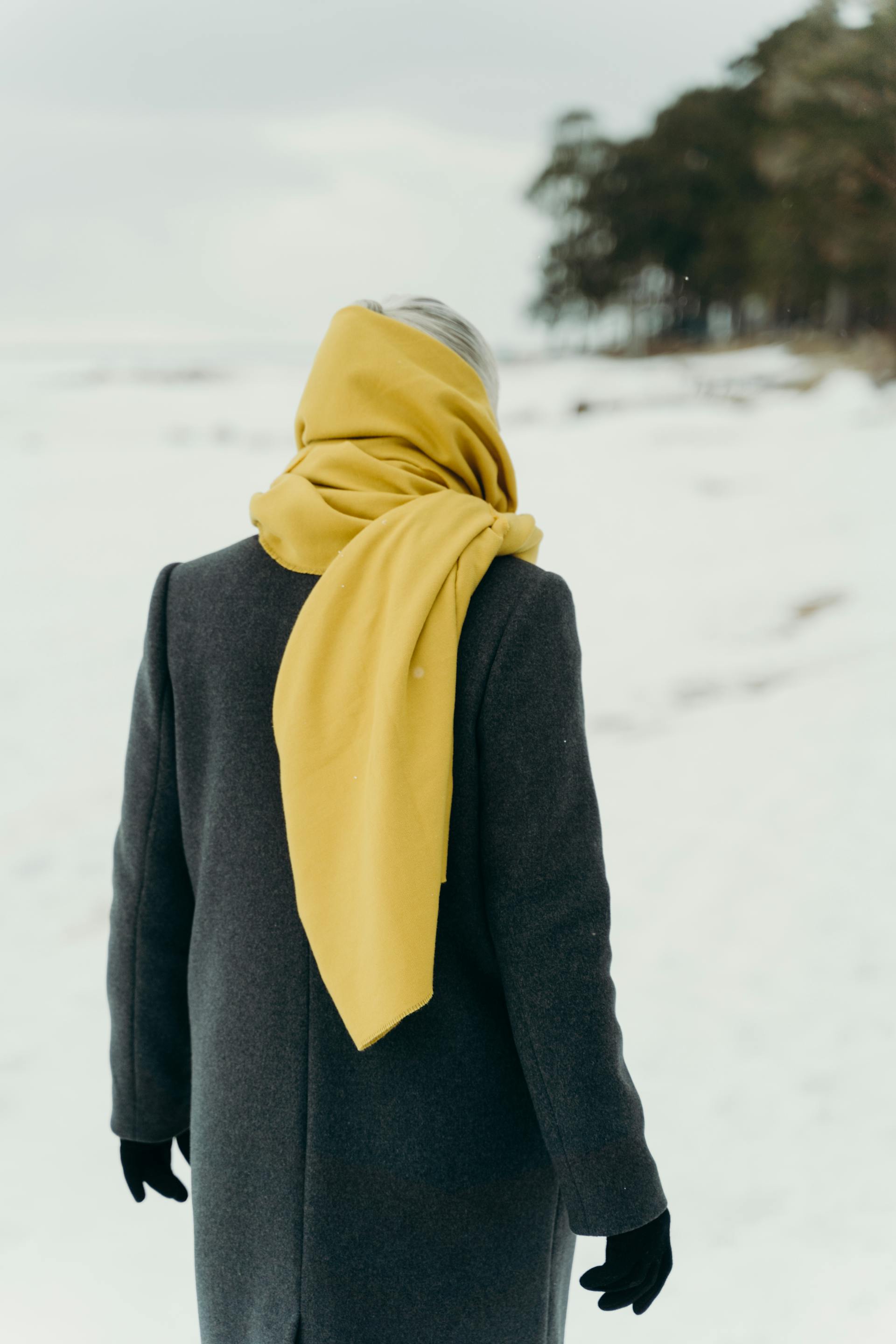 Une femme âgée vêtue d'un manteau et d'une écharpe jaune dans la neige, vue de dos | Source : Pexels