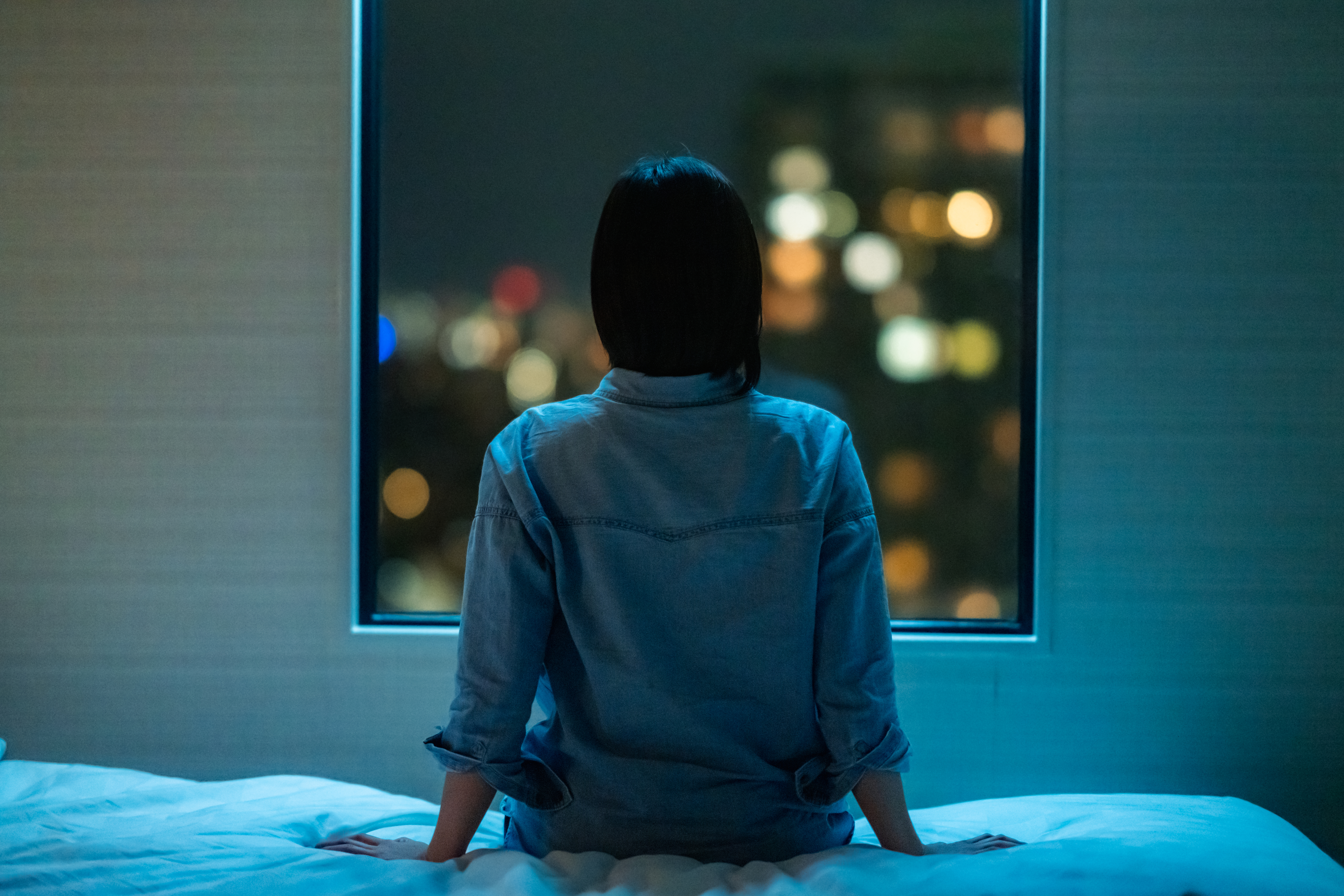 Vue arrière d'une femme assise seule sur un lit dans une chambre et regardant par la fenêtre la nuit | Source : Getty Images