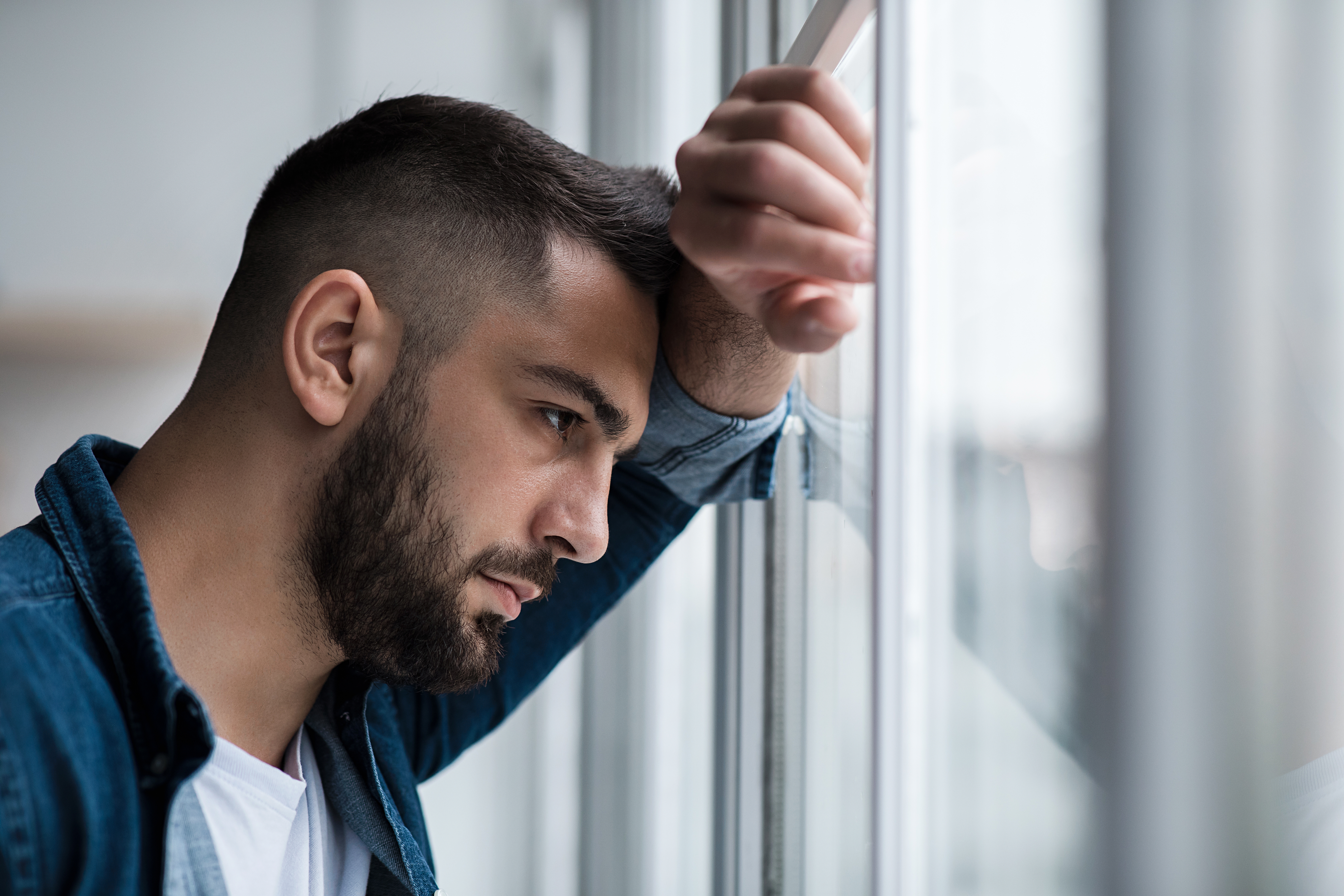 Hombre joven atractivo empanado infeliz mira por la ventana en el interior de un salón. | Fuente: Shutterstock