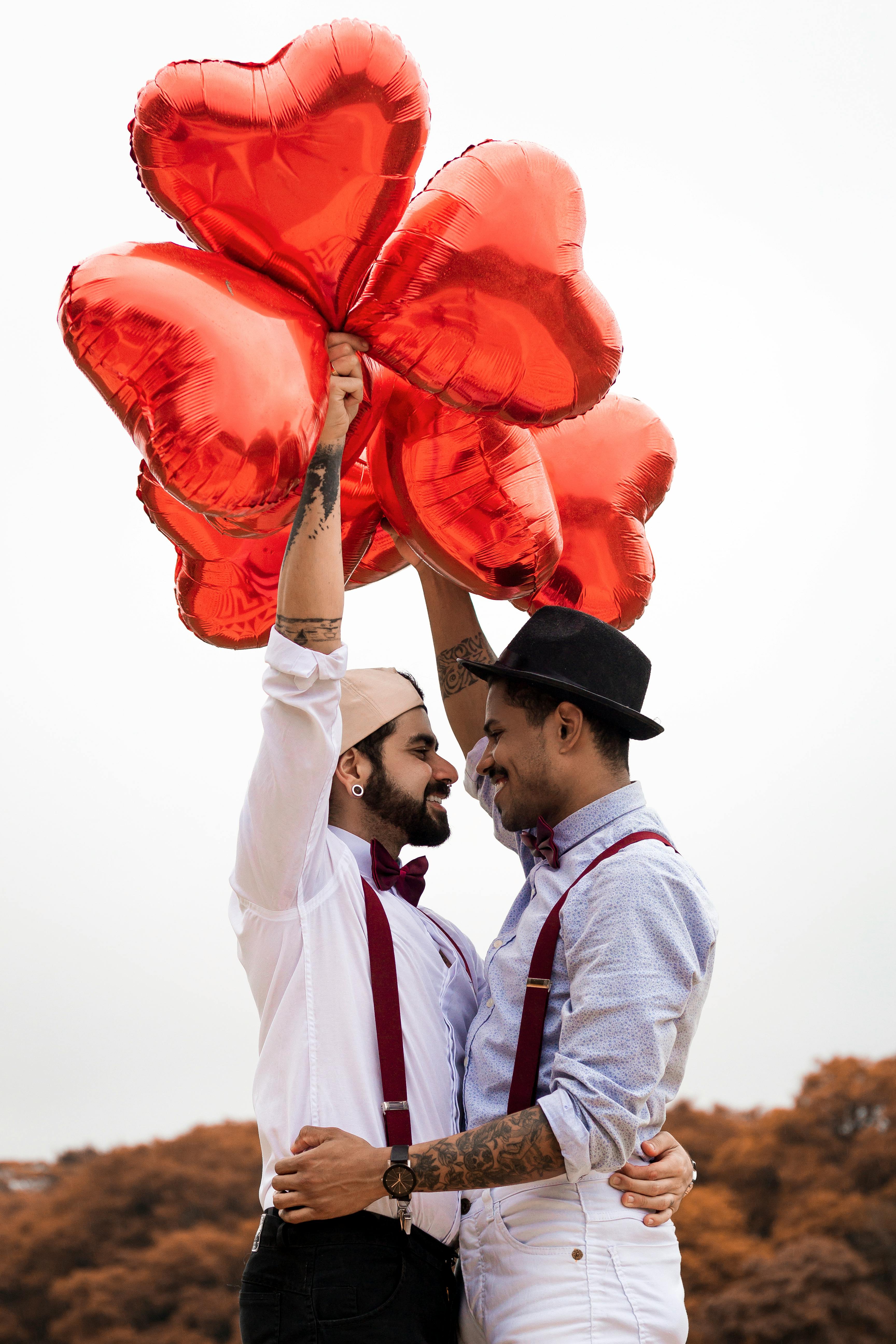 Deux hommes souriant et s'embrassant en brandissant des ballons en forme de cœur | Source : Pexels