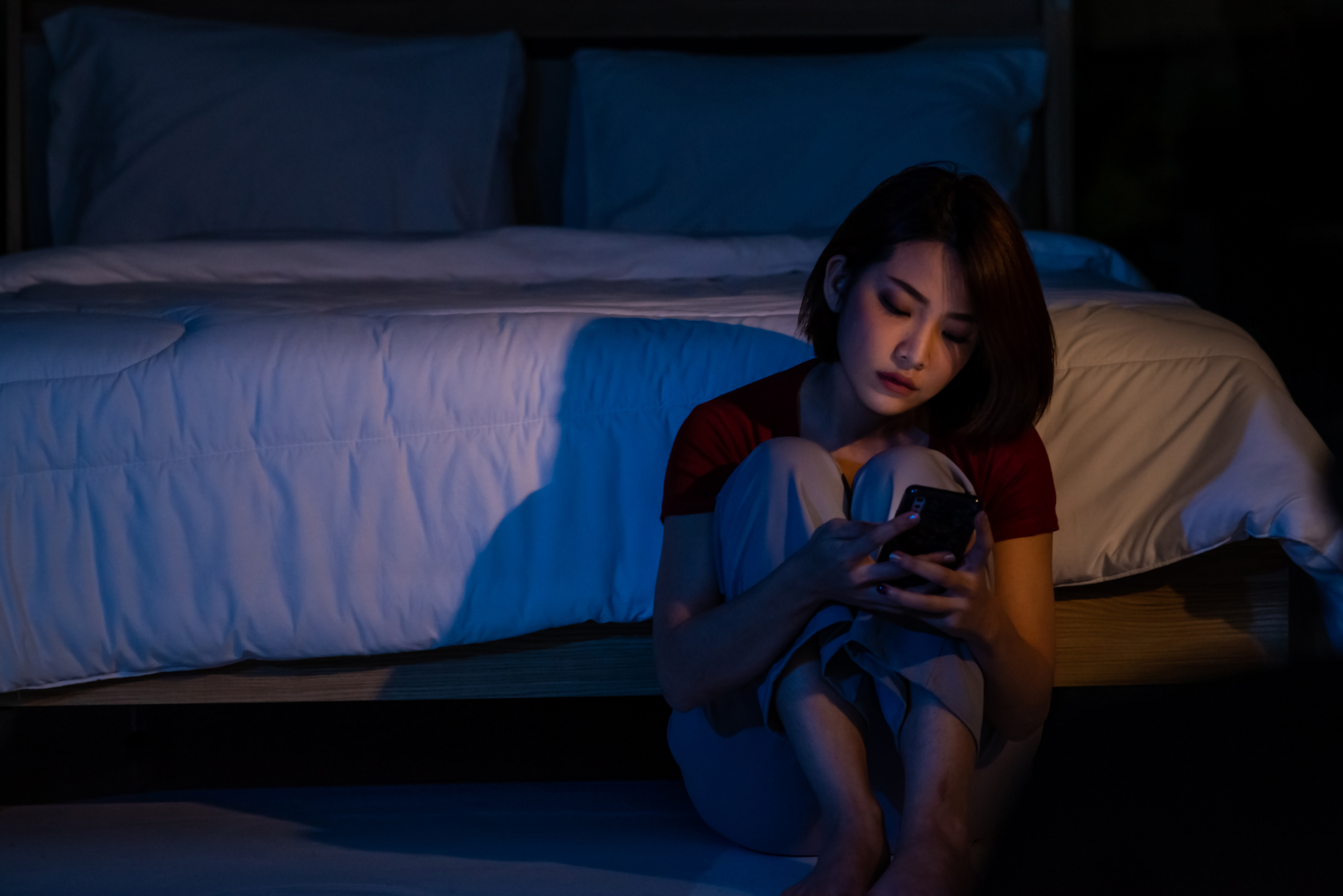 Jeune femme asiatique belle et déprimée assise seule sur le sol de la chambre en regardant son smartphone. | Source : Getty Images