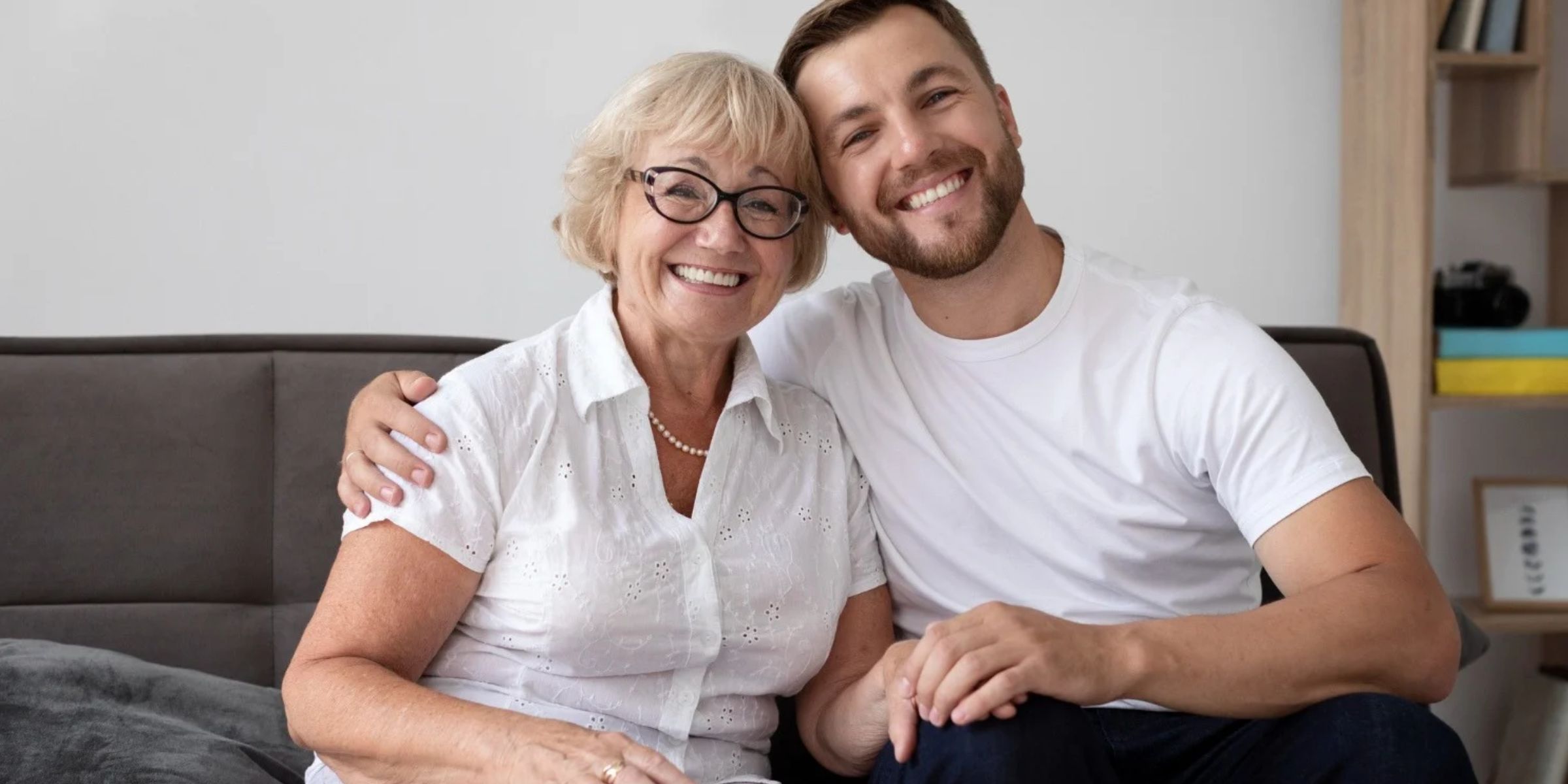 Une maman âgée qui sourit, assise sur un canapé avec son fils adulte, tous les deux enlacés | Source : Magnific
